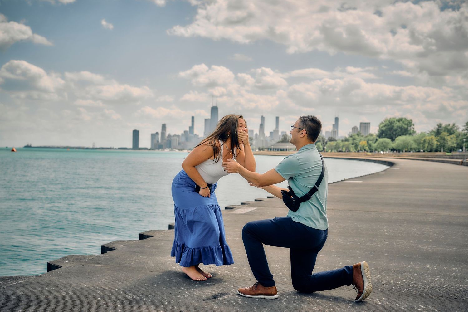 A proposal moment unfolds on a lakeside promenade with the Chicago skyline visible across the water on a sunny day.