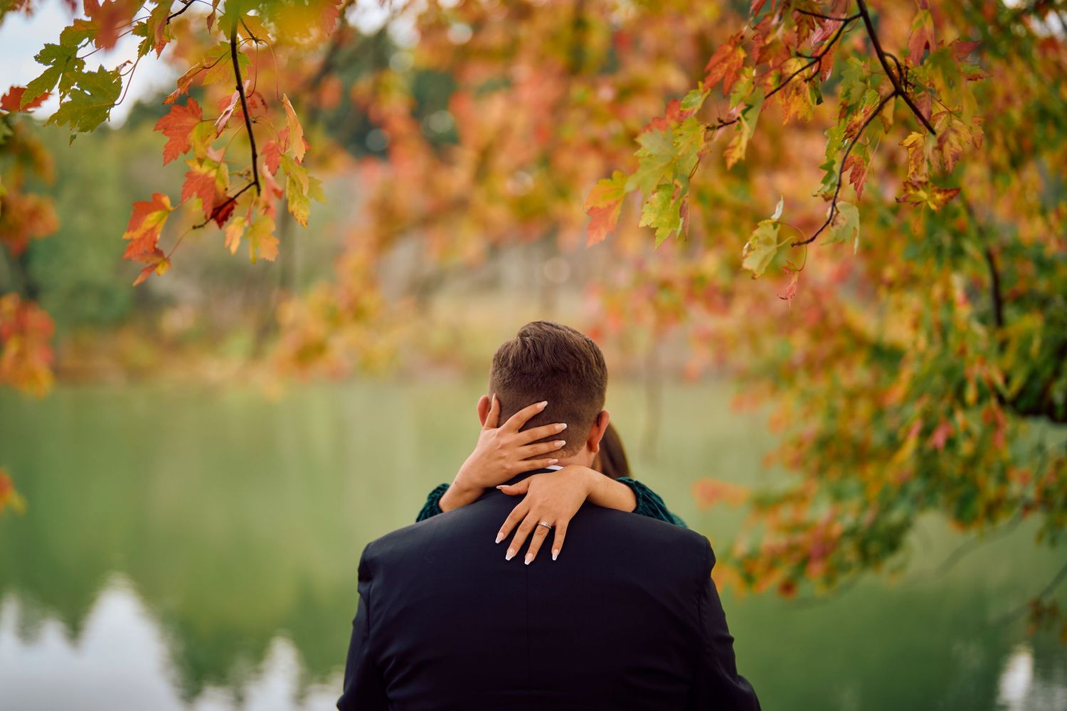 Couple embraces near a lake with autumn foliage and branches overhead during a romantic moment.