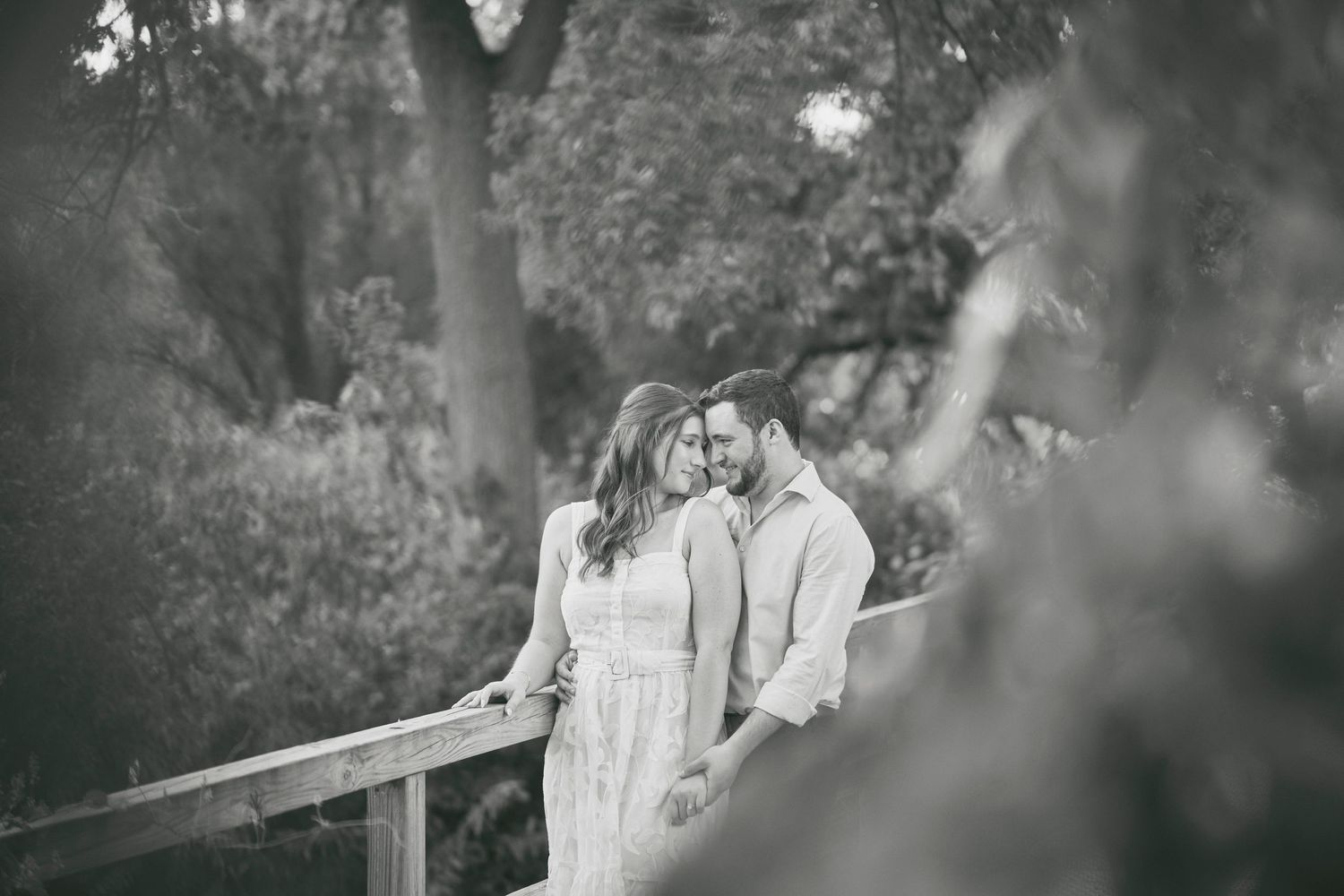 A romantic black and white portrait of a couple standing close together by a wooden railing in a garden setting.
