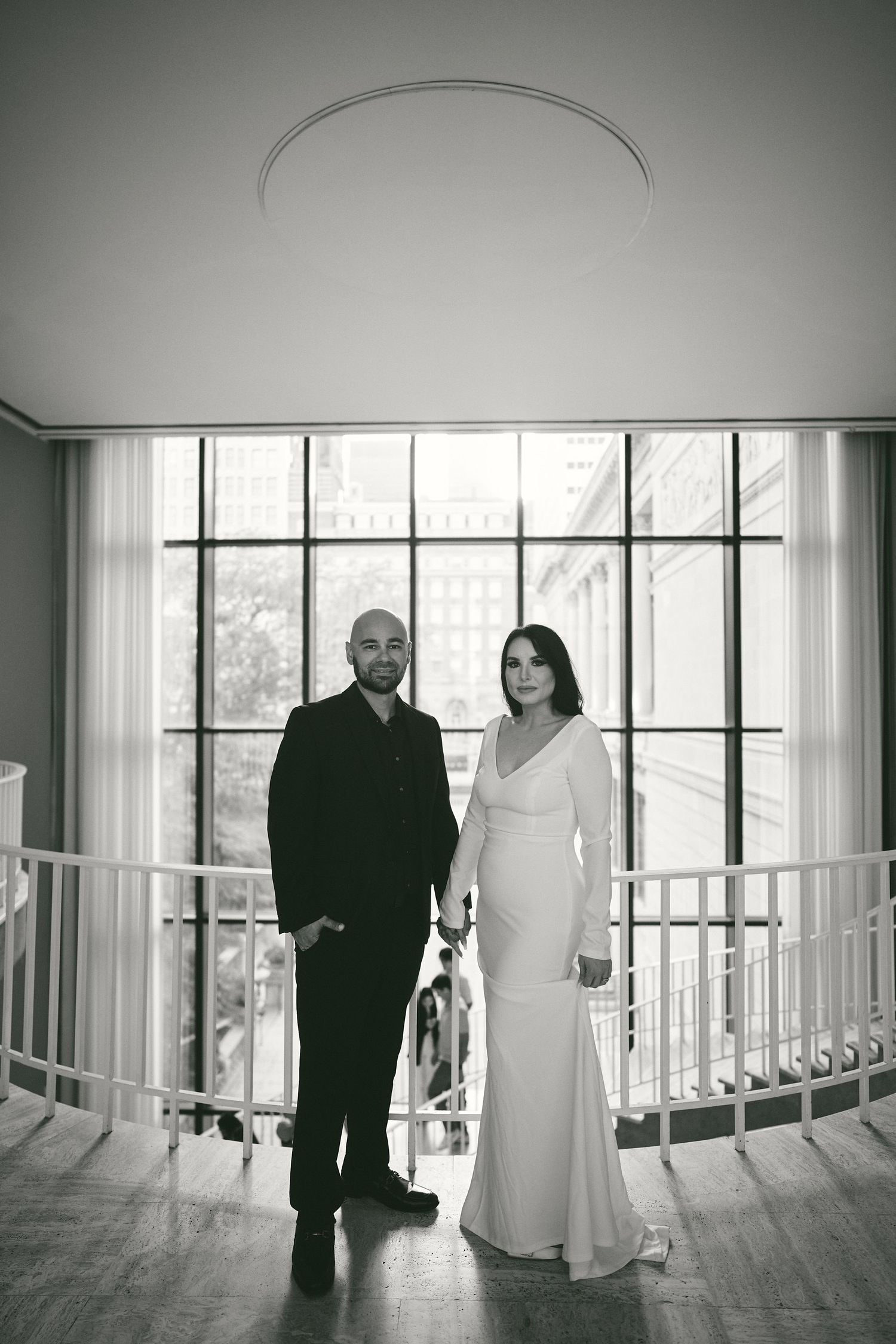 A couple stands elegantly in formal attire near large windows in a black and white wedding photograph.