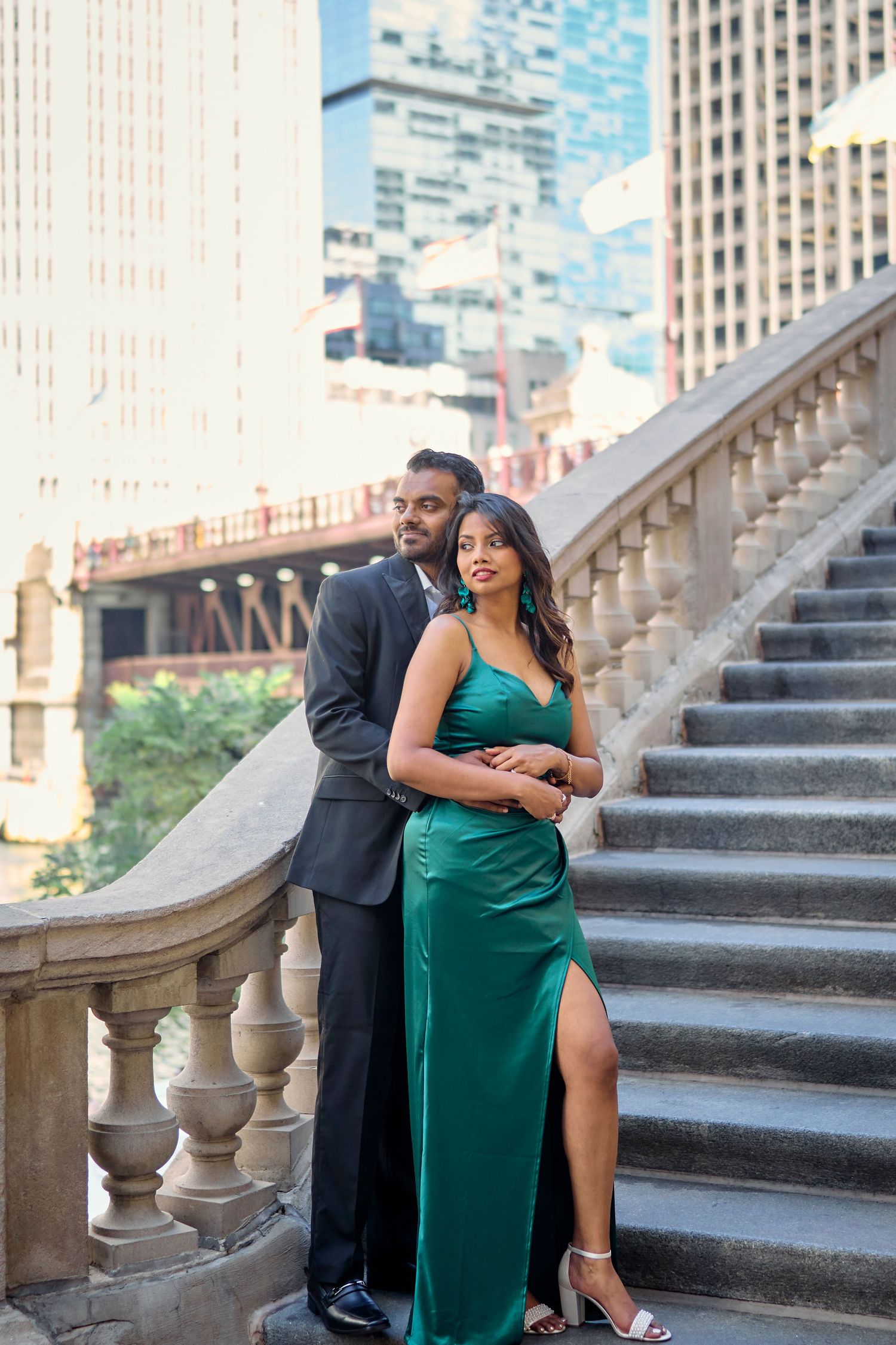 Elegantly dressed couple poses on grand stone staircase in downtown setting with city buildings in background.