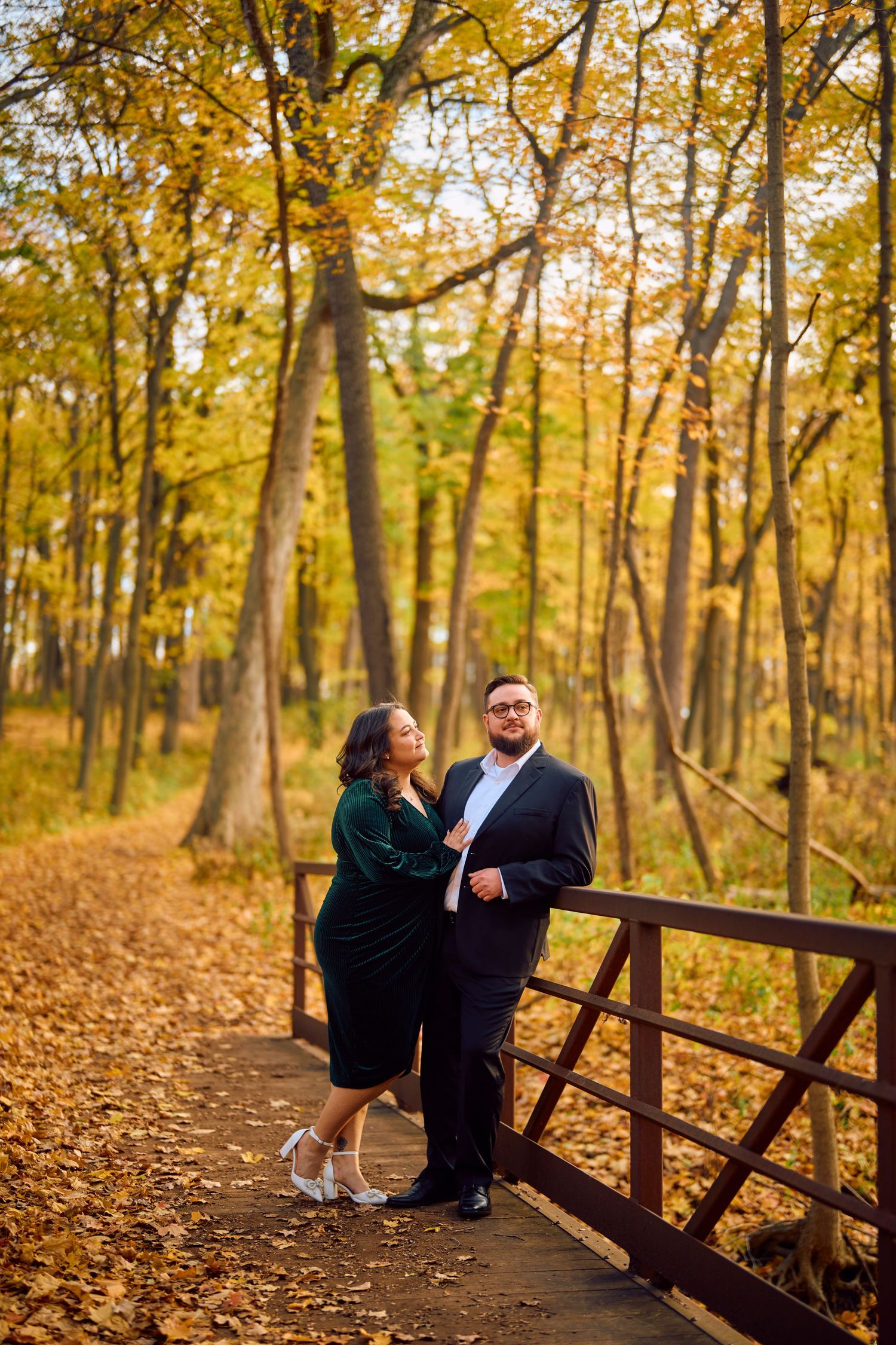 A couple stands together on a wooden bridge surrounded by golden autumn foliage in a forest setting.