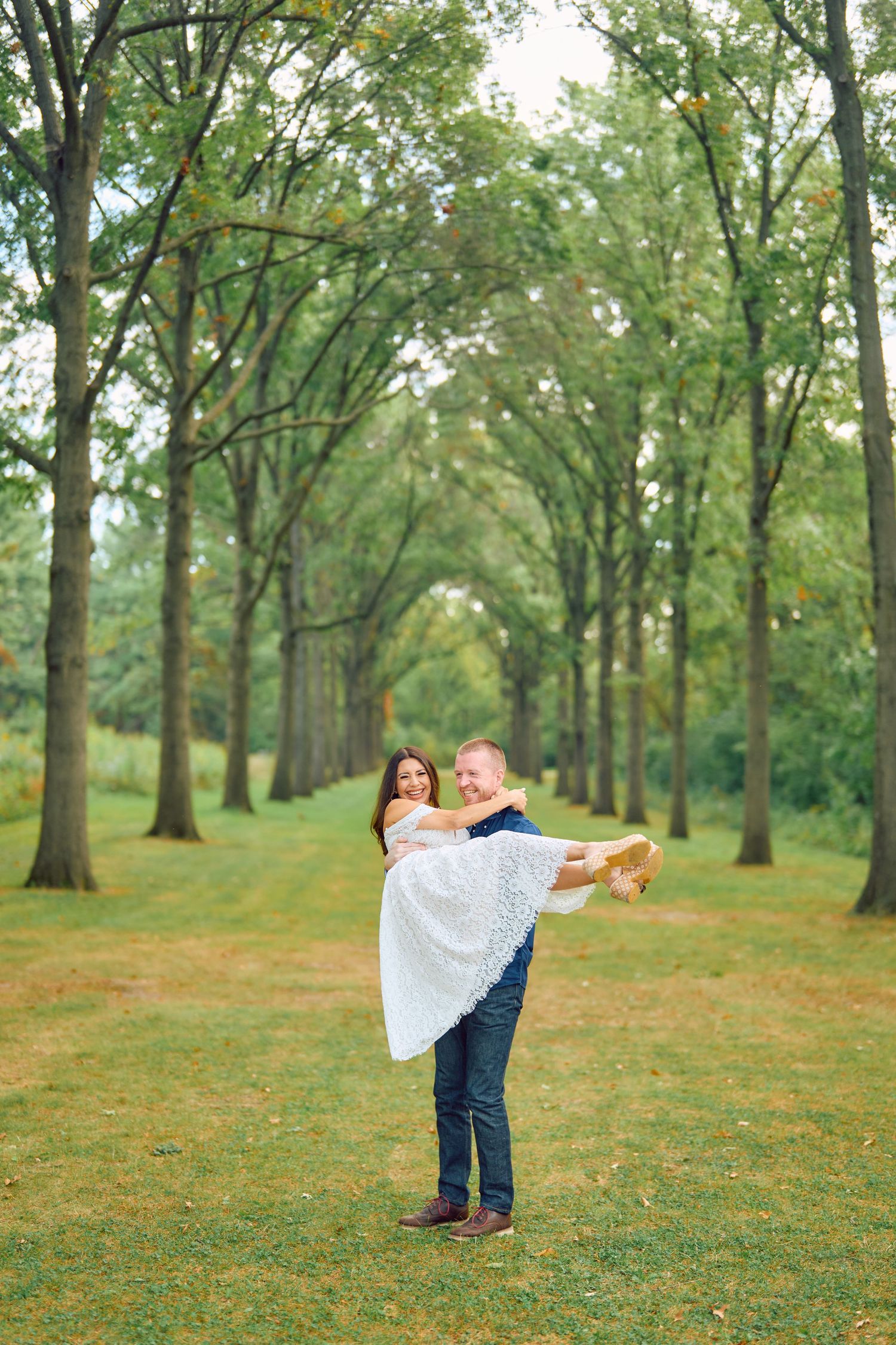 A romantic couple embraces on a tree-lined path with tall green trees forming a natural archway overhead.