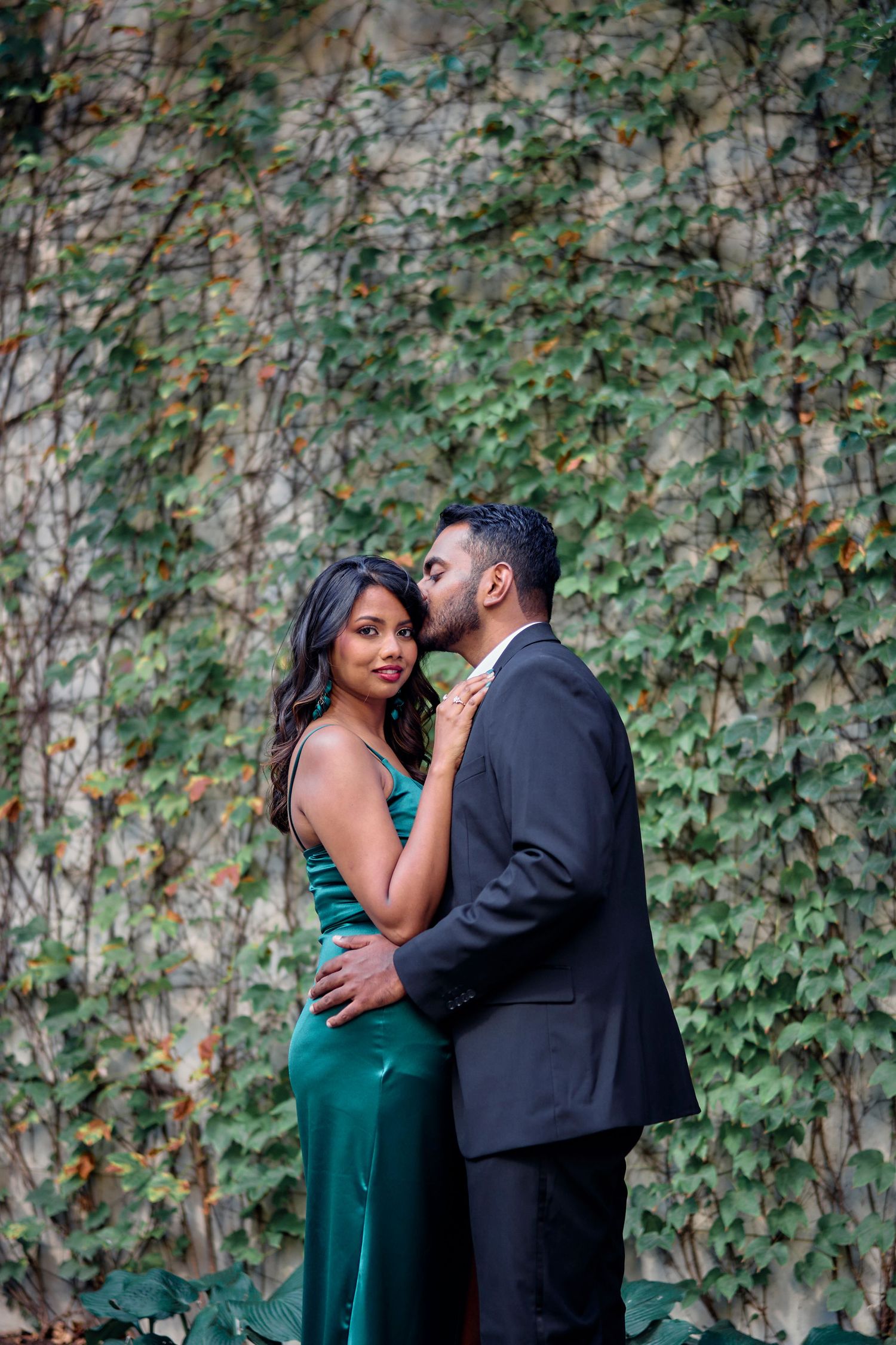 A couple in formalwear embrace against an ivy-covered wall backdrop.