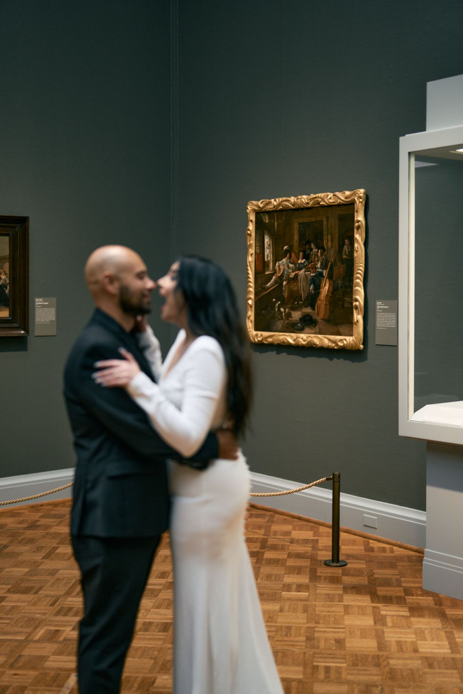 Romantic couple shares an intimate moment in an art museum gallery with classic paintings and parquet flooring.