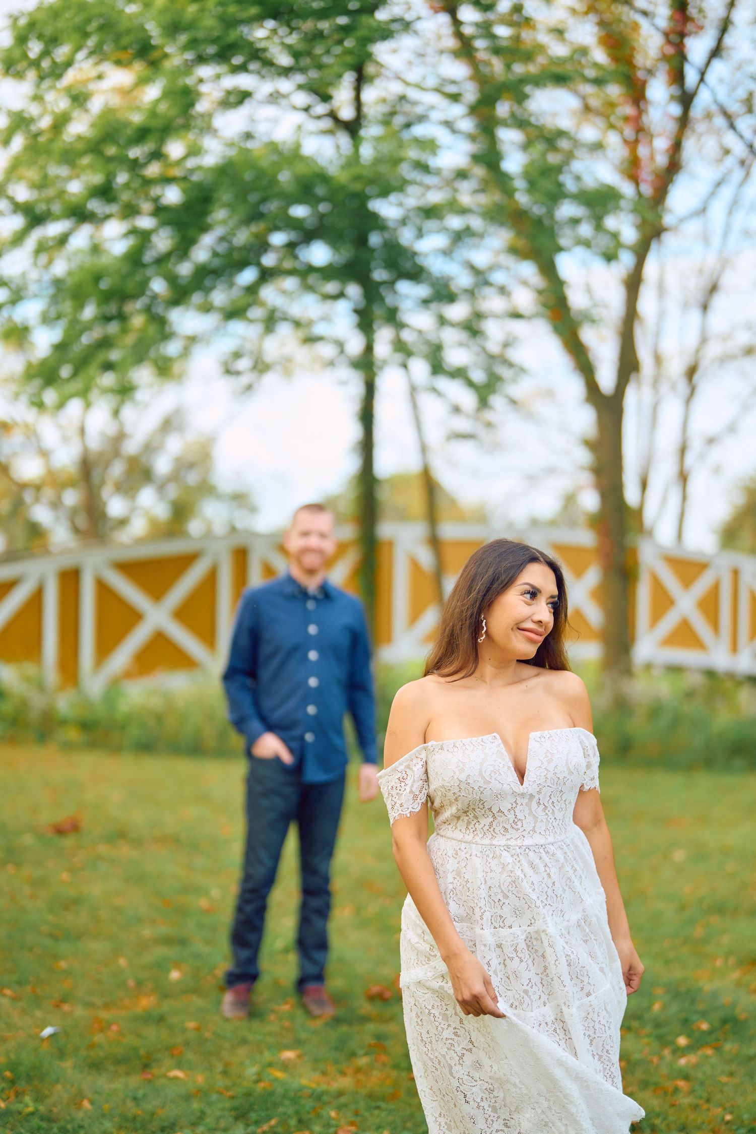 A couple poses outdoors in a garden setting with white fence behind them on a sunny autumn day.