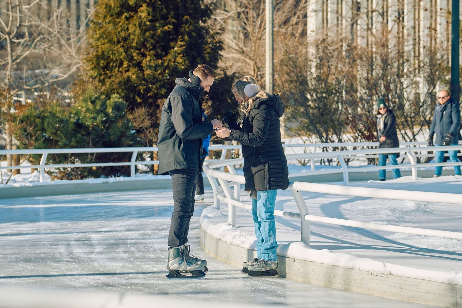 Two people ice skating together on an outdoor rink in winter surrounded by snow and evergreen trees.