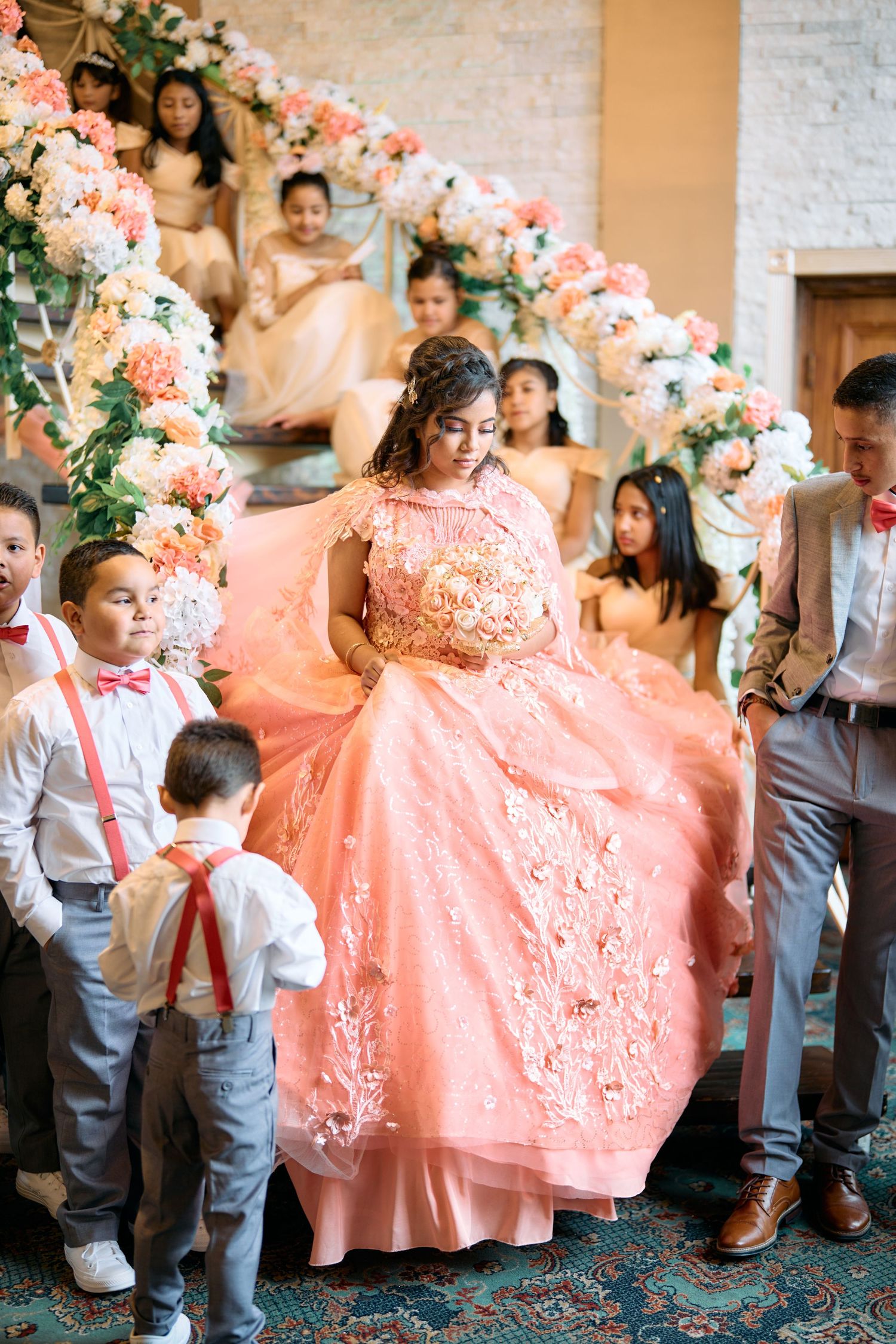 A quinceañera celebration with someone in a flowing coral pink ballgown surrounded by floral arrangements and guests.