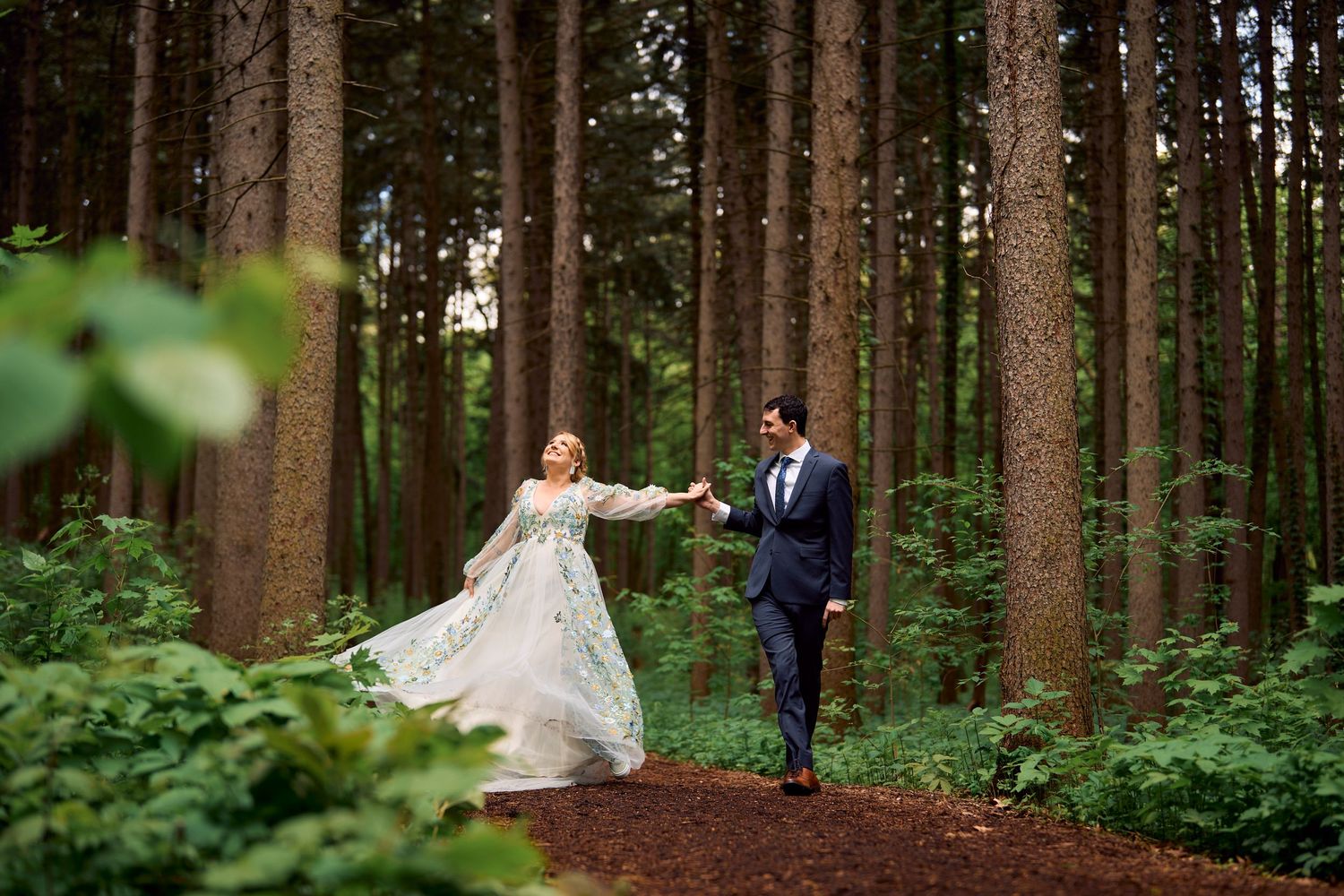 A couple walks hand in hand through a lush forest path, the bride wearing a flowing lace gown among tall pine trees.