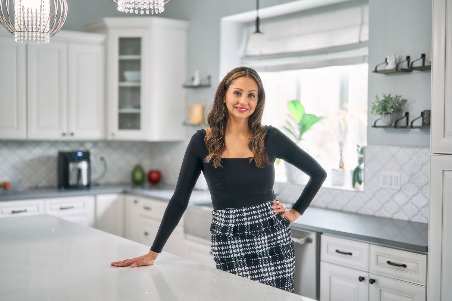 Woman in black top and plaid skirt leaning on white kitchen counter.
