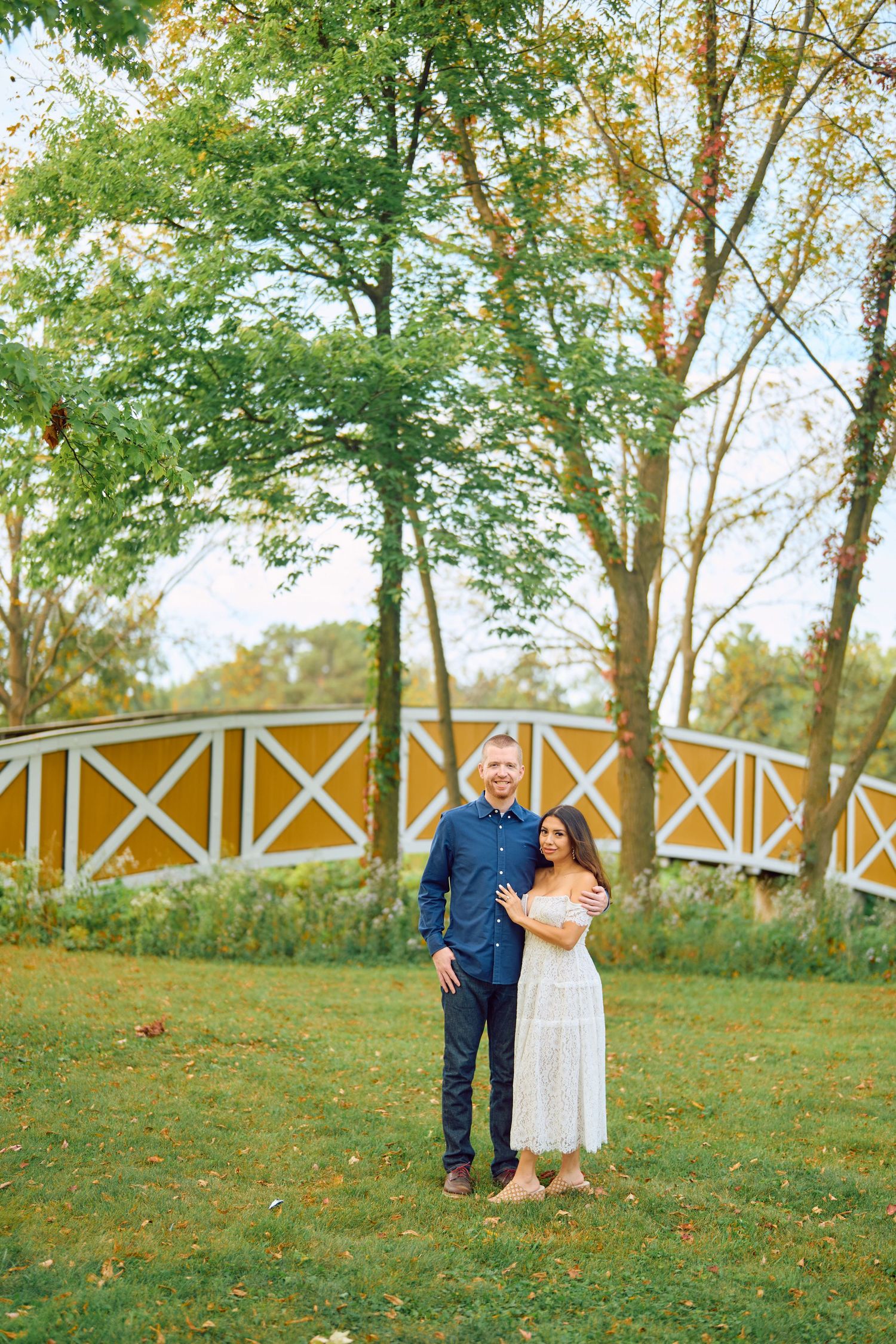 A couple stands together in front of a white and yellow fence on grassy grounds with tall trees in the background.