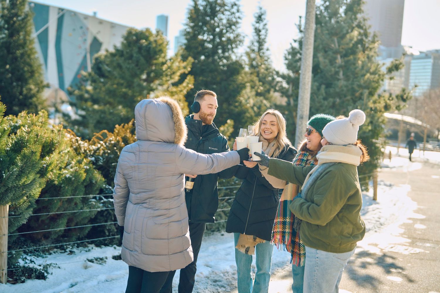 A group of people laughing together outdoors in winter with snow on the ground and evergreen shrubs nearby.