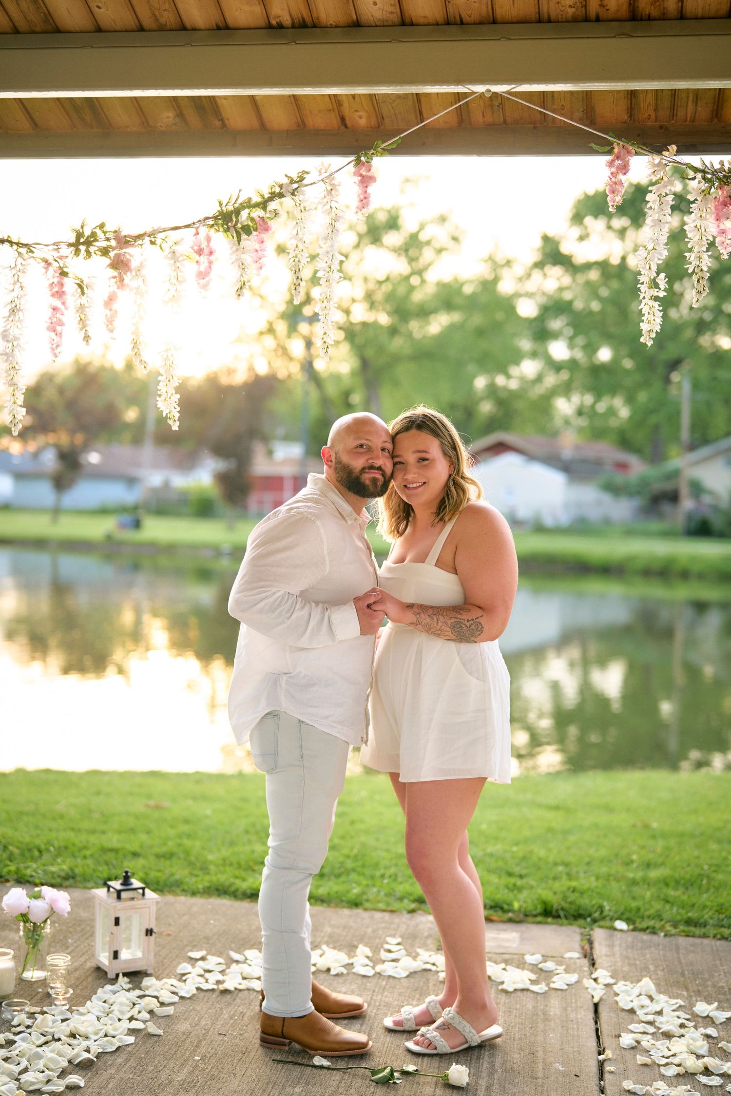 A couple embraces on a lakeside deck decorated with hanging floral garlands during golden hour sunset.