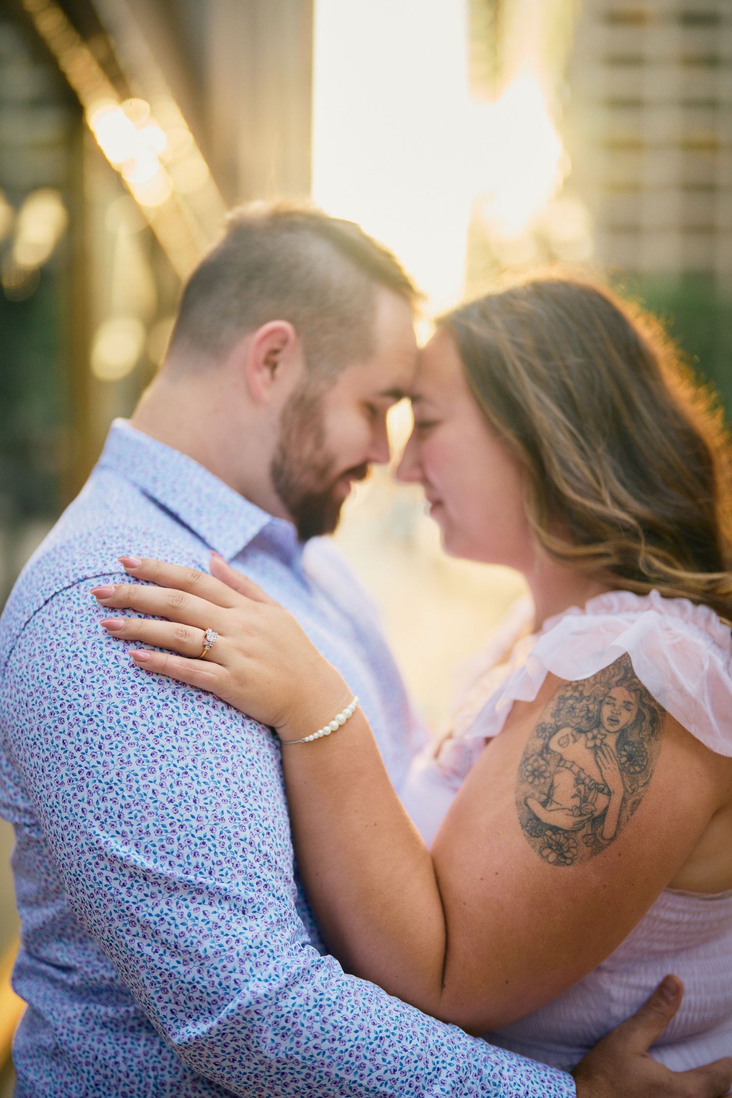 A romantic couple embraces in soft sunlight, with the woman's arm showing a detailed tattoo on her upper arm.