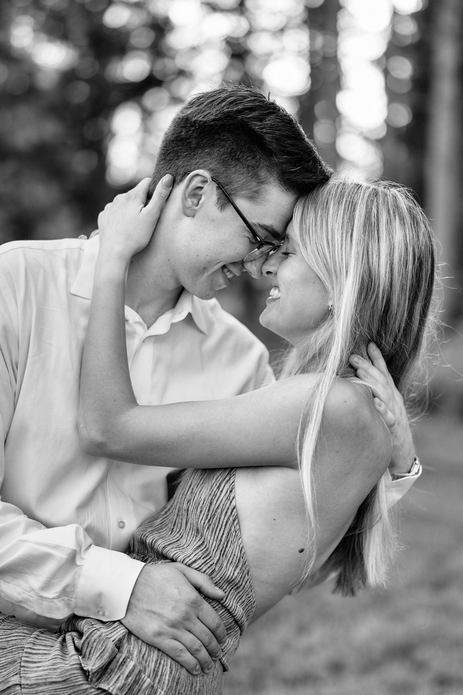 A romantic black and white portrait of a couple embracing and smiling together in a forest setting.
