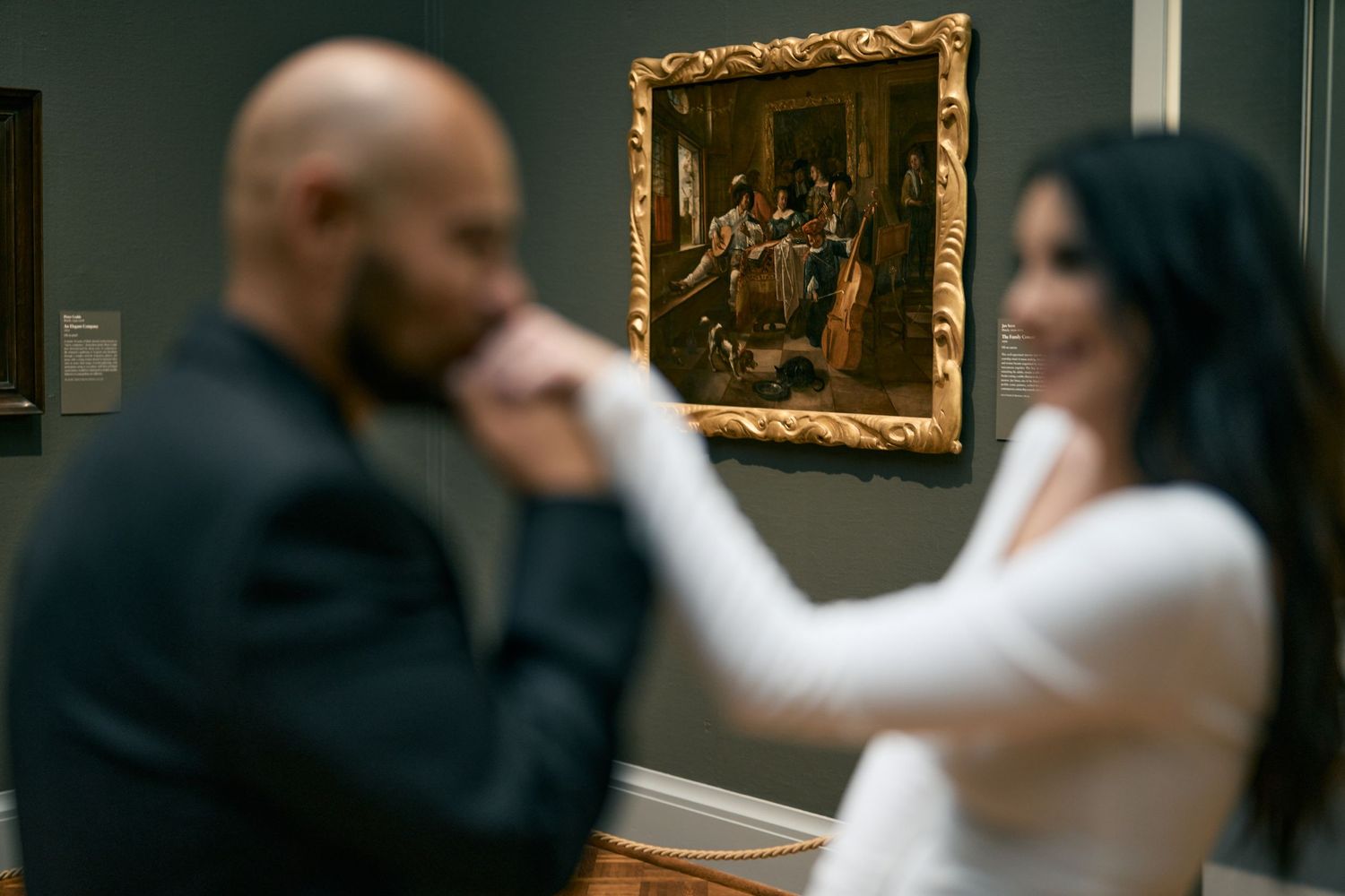 Two people stand in an art gallery looking at a framed classical painting mounted on a dark wall.