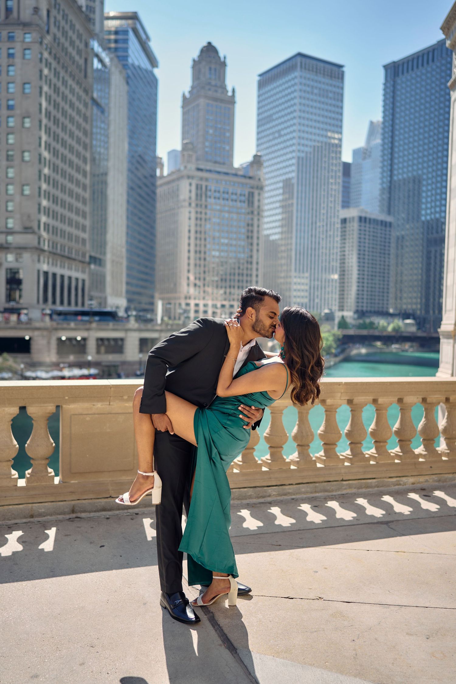 A romantic couple embraces on a balcony overlooking the Chicago River with the downtown skyline visible in the background.