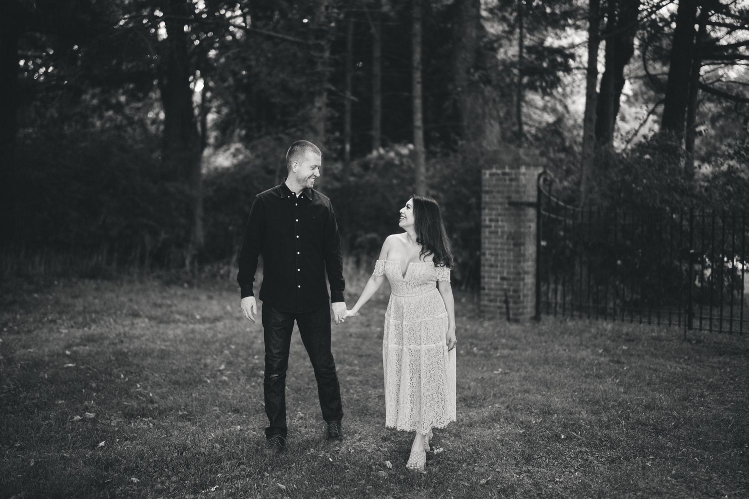 A couple stands together in a black and white photo on a grassy area near a garden gate.