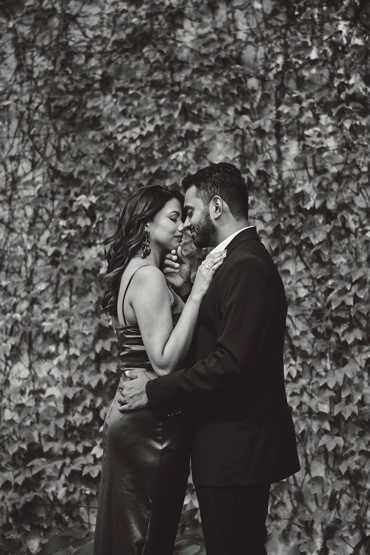 A romantic black and white photo of a couple embracing against an ivy-covered wall.