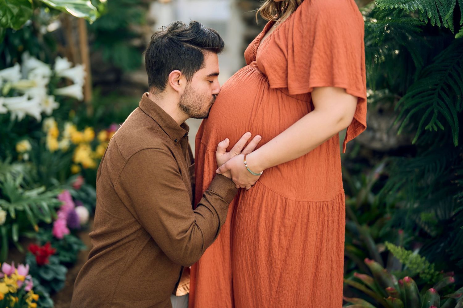 A tender moment captured in a garden as a person in brown embraces a pregnant figure wearing a flowing orange dress.