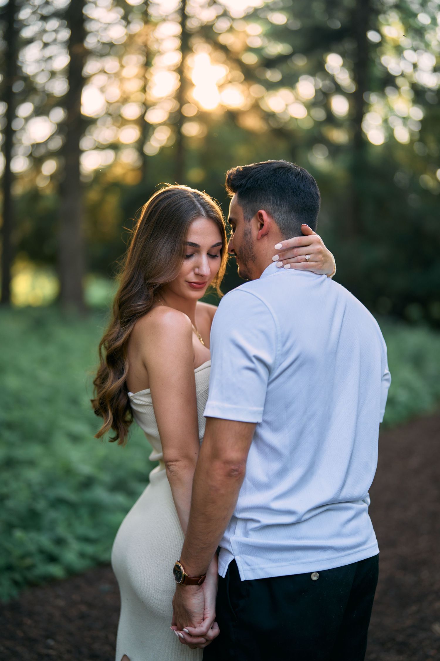 A romantic couple embraces in an outdoor setting during golden hour among trees.