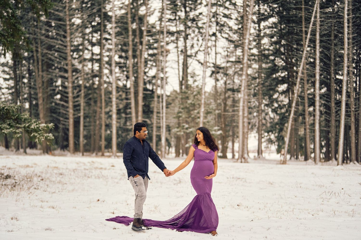A romantic couple holding hands in a snowy forest setting with a flowing purple dress creating an elegant winter scene.