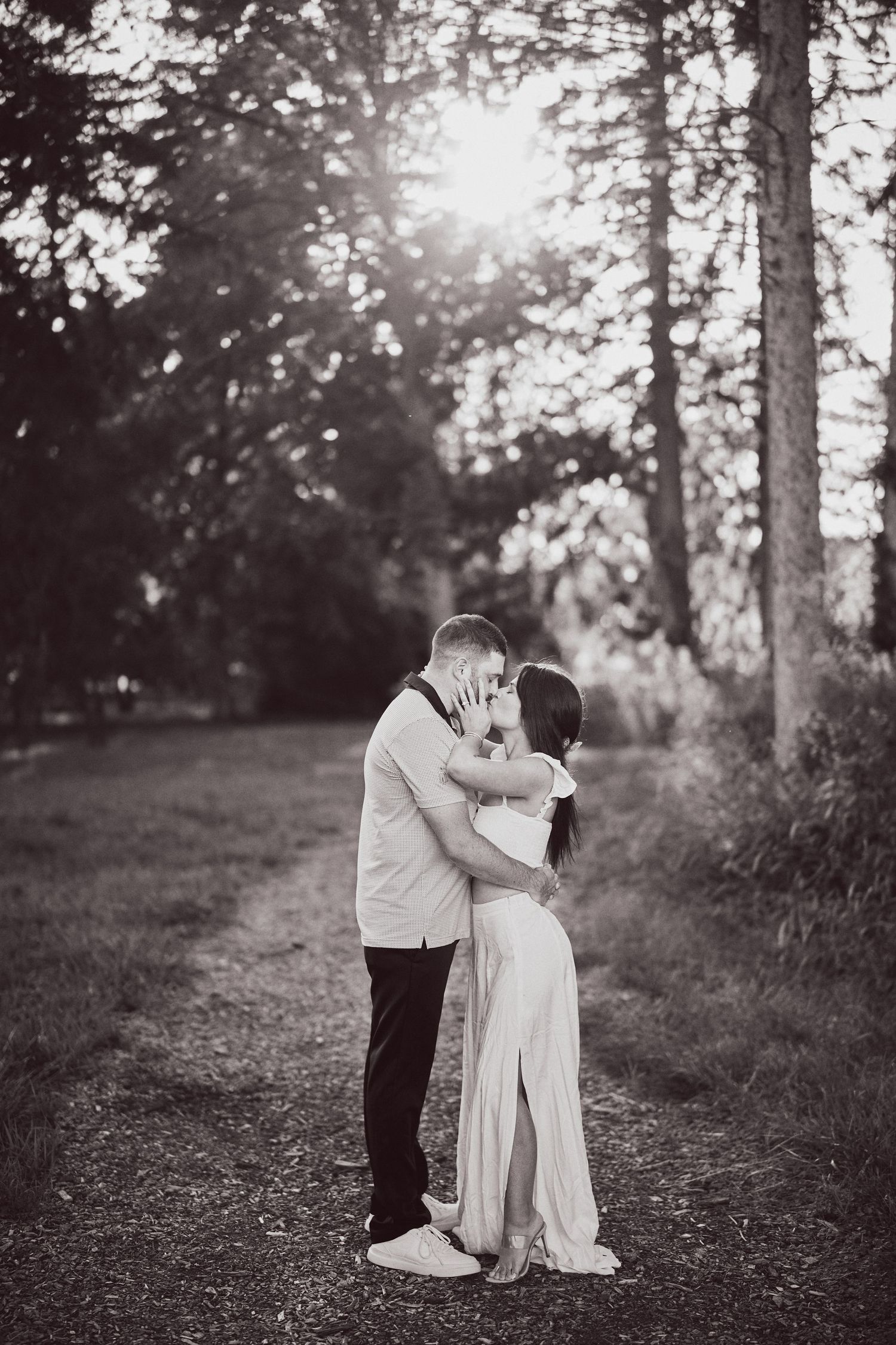 A couple shares an intimate moment on a sunlit forest path in black and white photography.