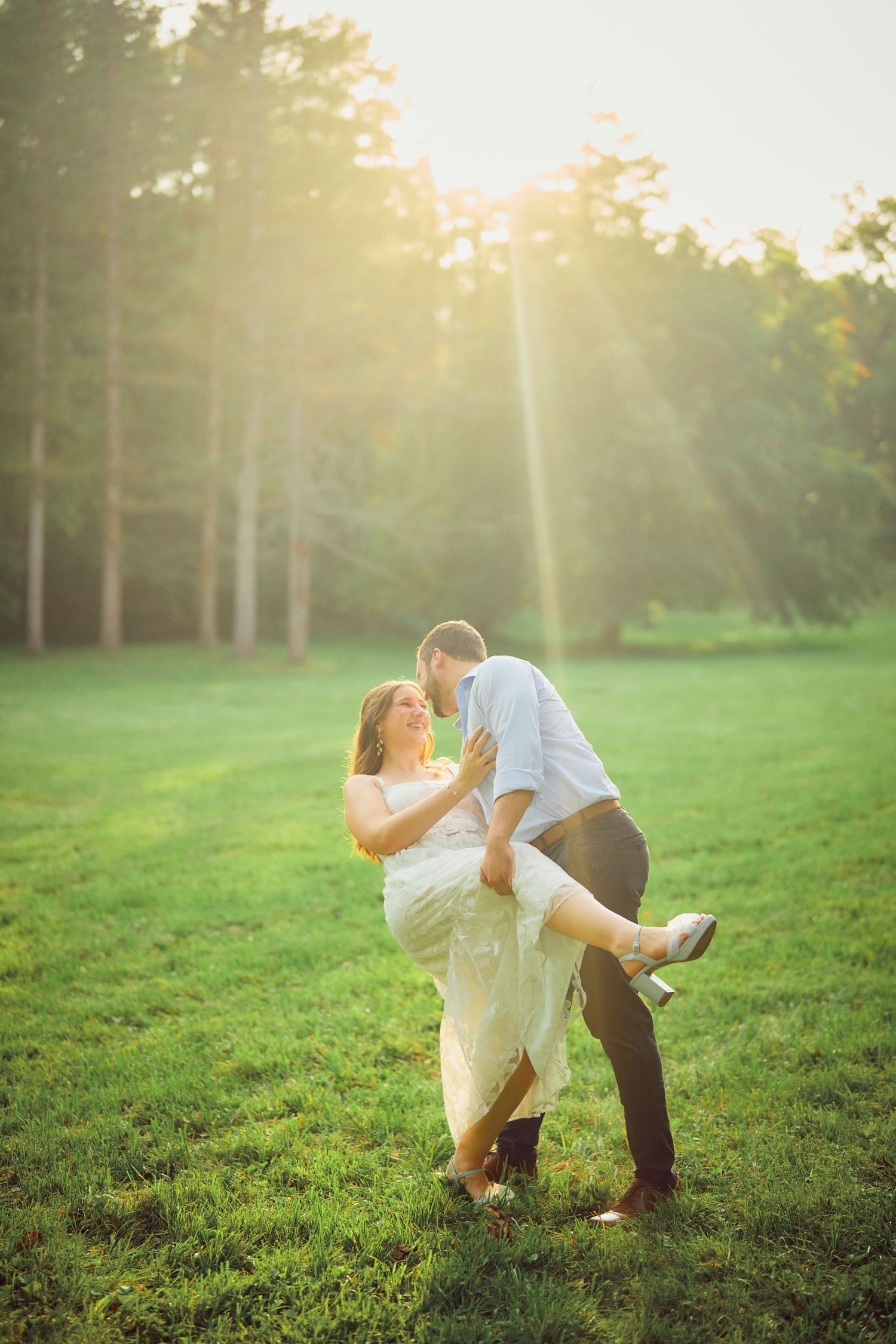 A romantic sunset engagement photo session in a grassy meadow with sun rays streaming through trees in the background.