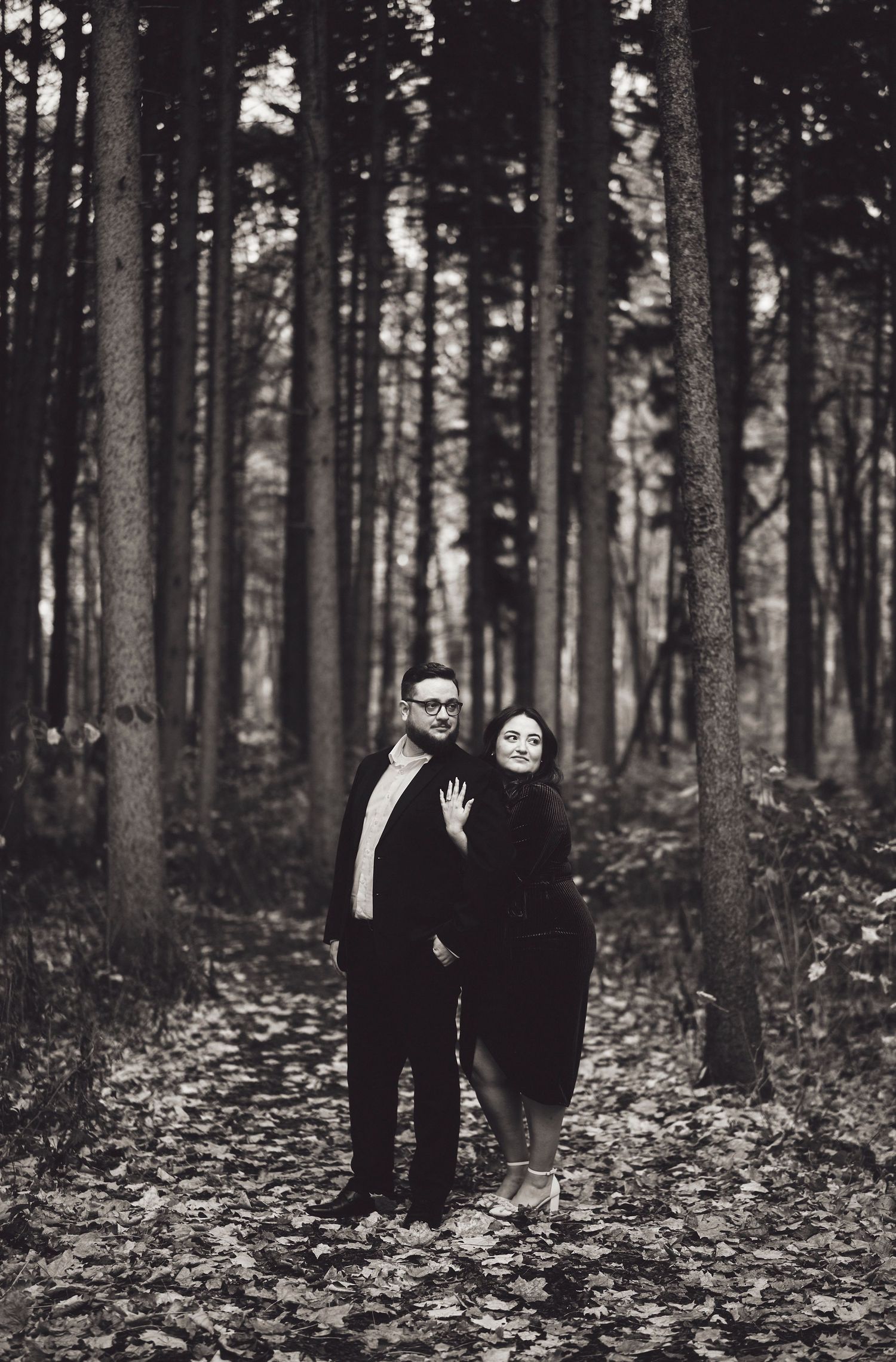 A couple standing together in a moody black and white forest setting surrounded by tall trees and fallen leaves.