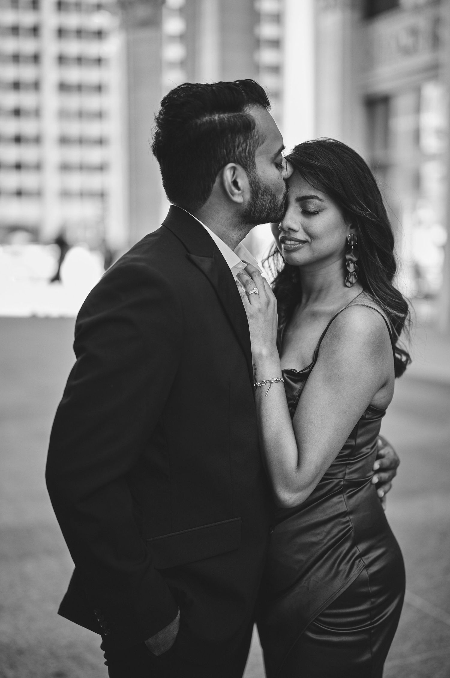 A romantic black and white portrait of a couple embracing intimately in an urban setting with tall buildings behind them.