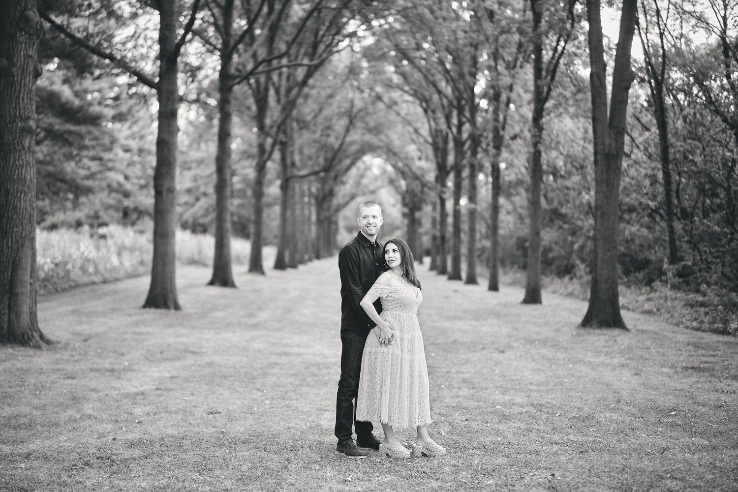 A romantic couple embraces on a tree-lined path in black and white photography.
