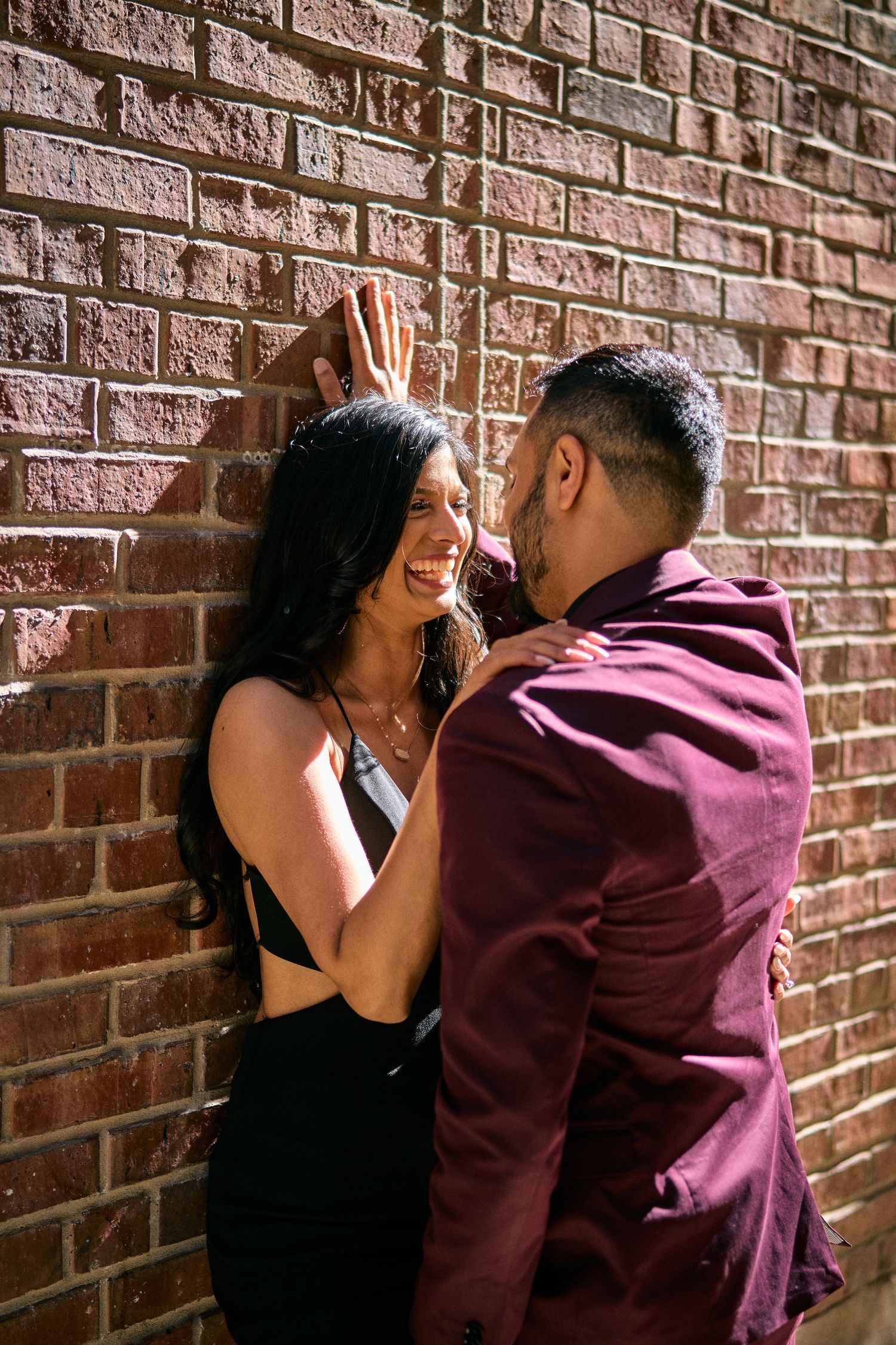 A couple sharing a romantic moment against a brick wall, she in black dress and he in burgundy jacket.