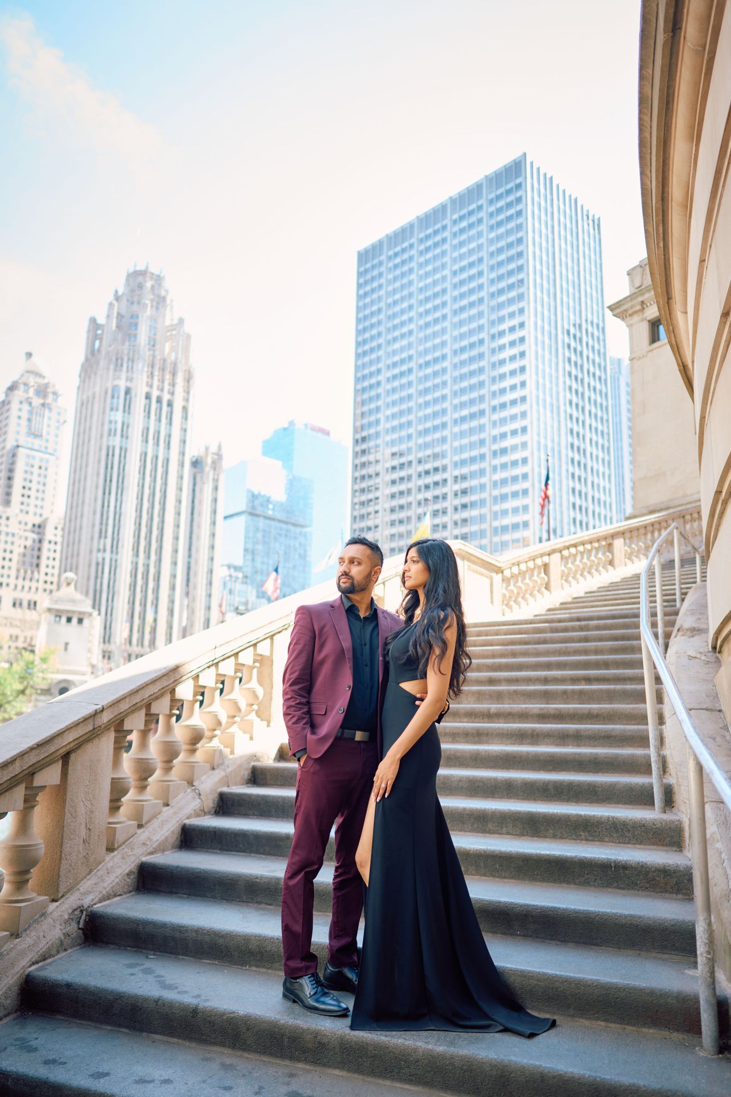 Couple in formal attire standing on grand stone staircase with Chicago skyscrapers in background.