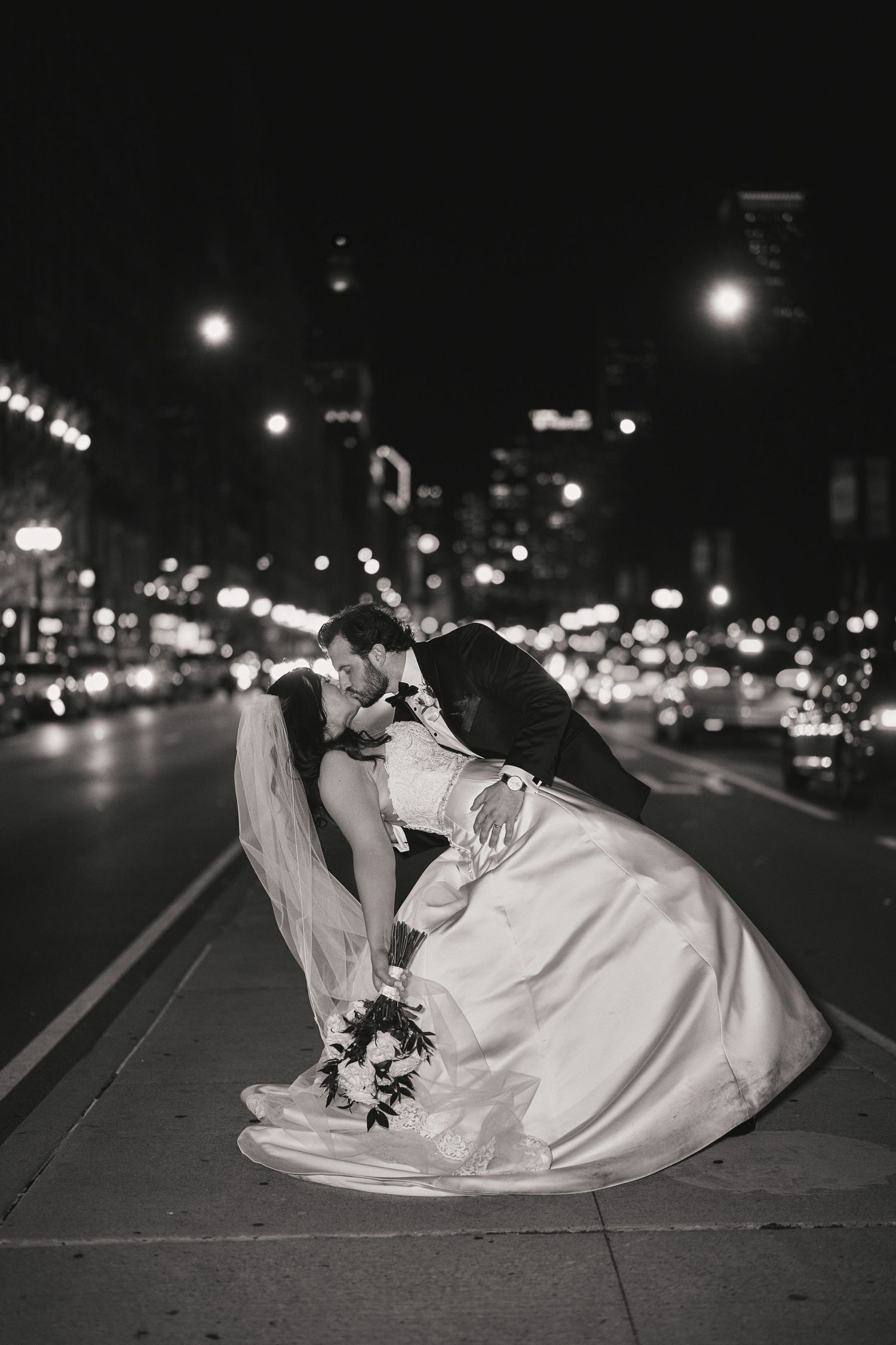 A romantic black and white cityscape wedding photo showing a couple embracing on a street at night with glowing lights.