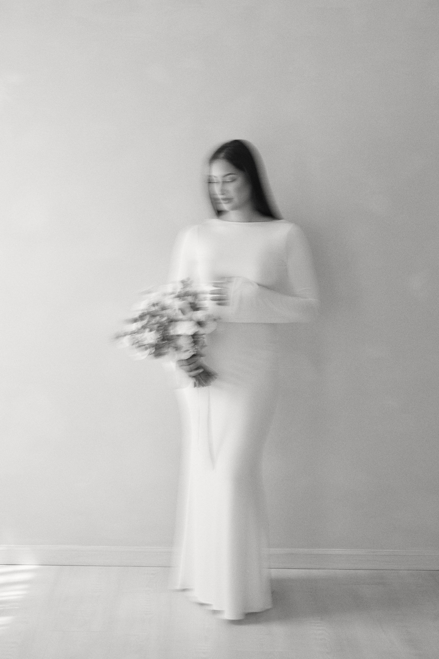 Ethereal black and white portrait of figure in flowing white dress holding flowers against minimal background.