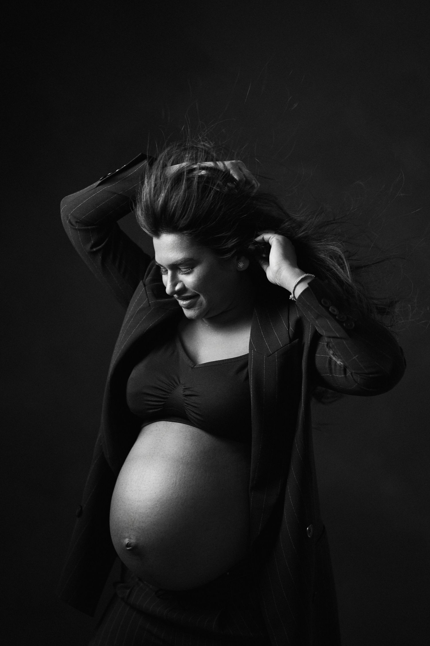 Artistic black and white maternity portrait showing an expectant mother in a dark cardigan against a dark background.