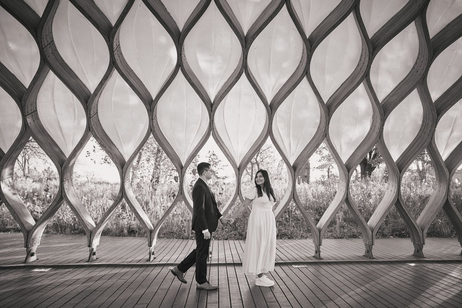 Two people in dark and light clothing stand on a wooden deck against a patterned diamond lattice architectural feature.