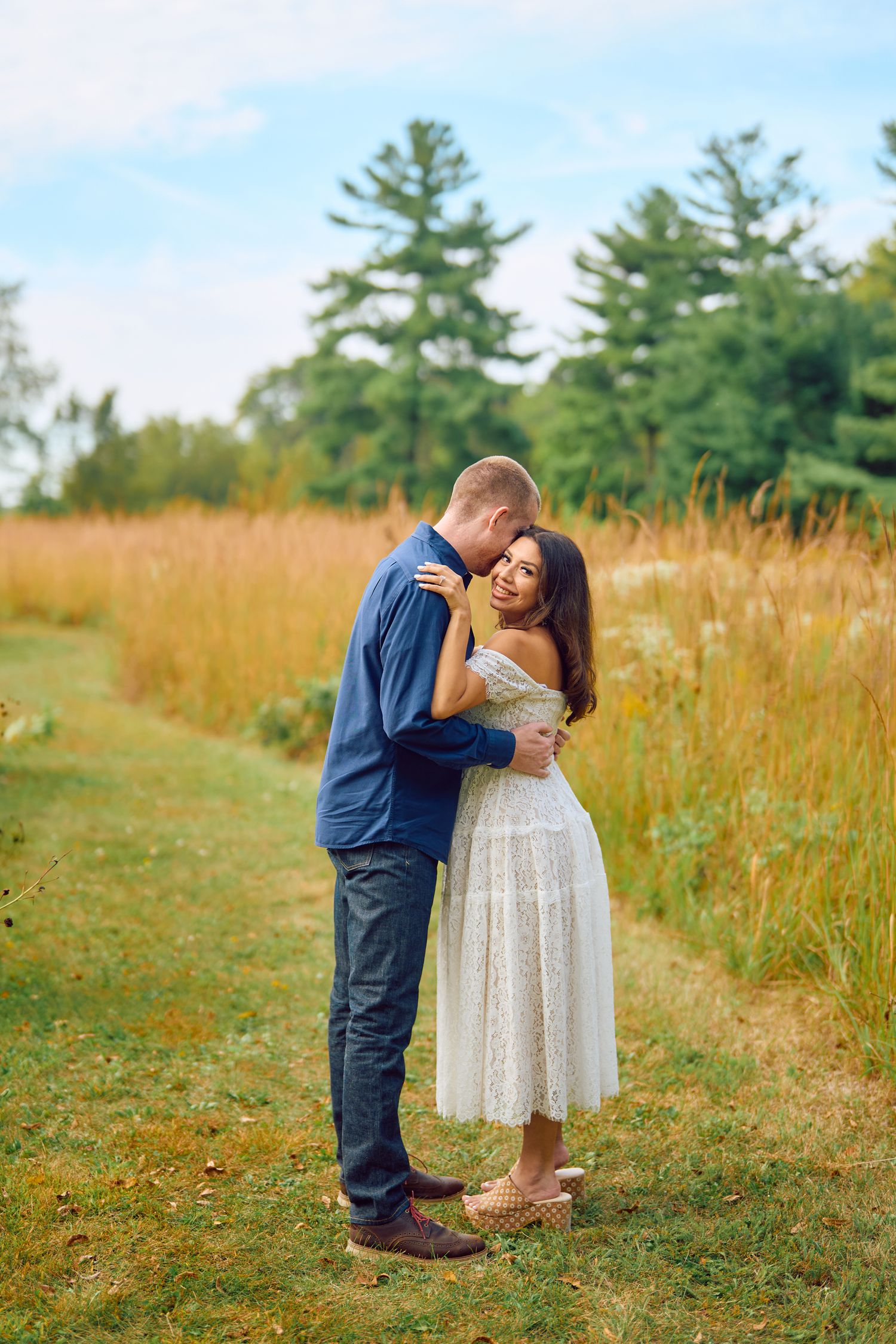A tender embrace between two people standing on a grassy path with tall autumn grasses and pine trees in the background.
