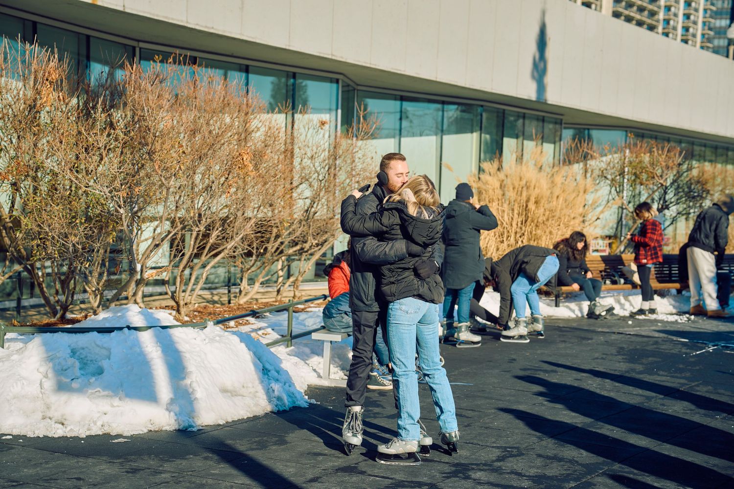 People gather outside a building on a sunny winter day with patches of snow and dormant shrubs in the background.