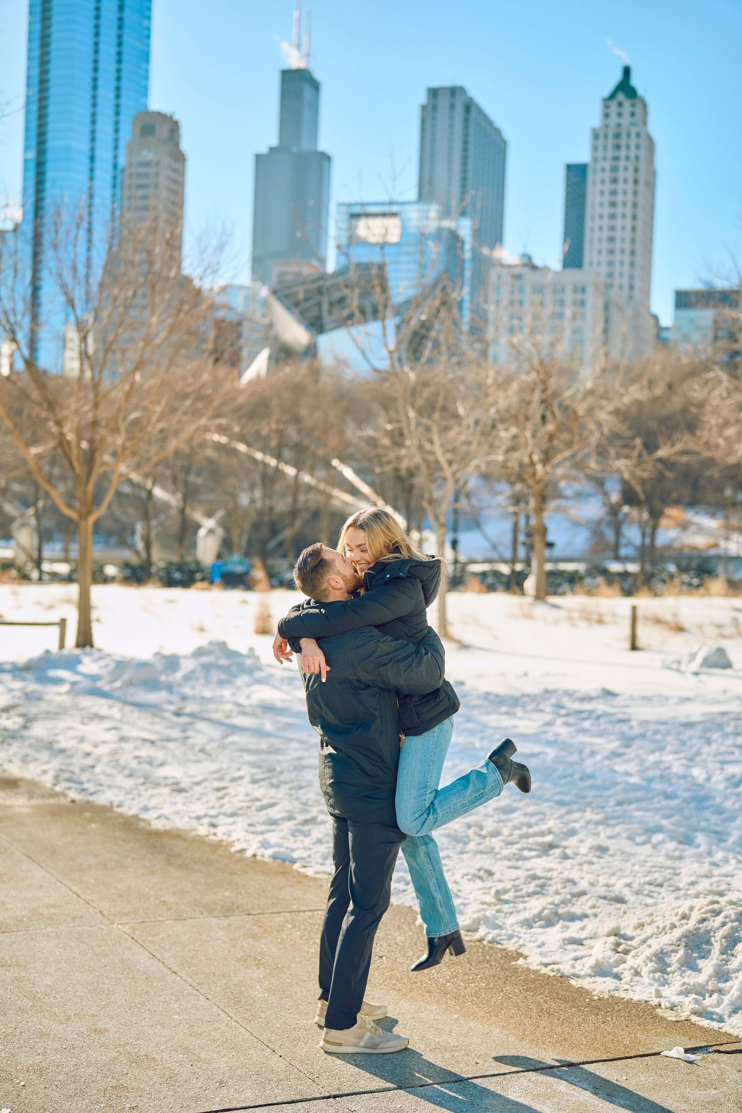 A couple embraces in winter at Chicago's downtown skyline with Willis Tower and snow-covered ground in the background.