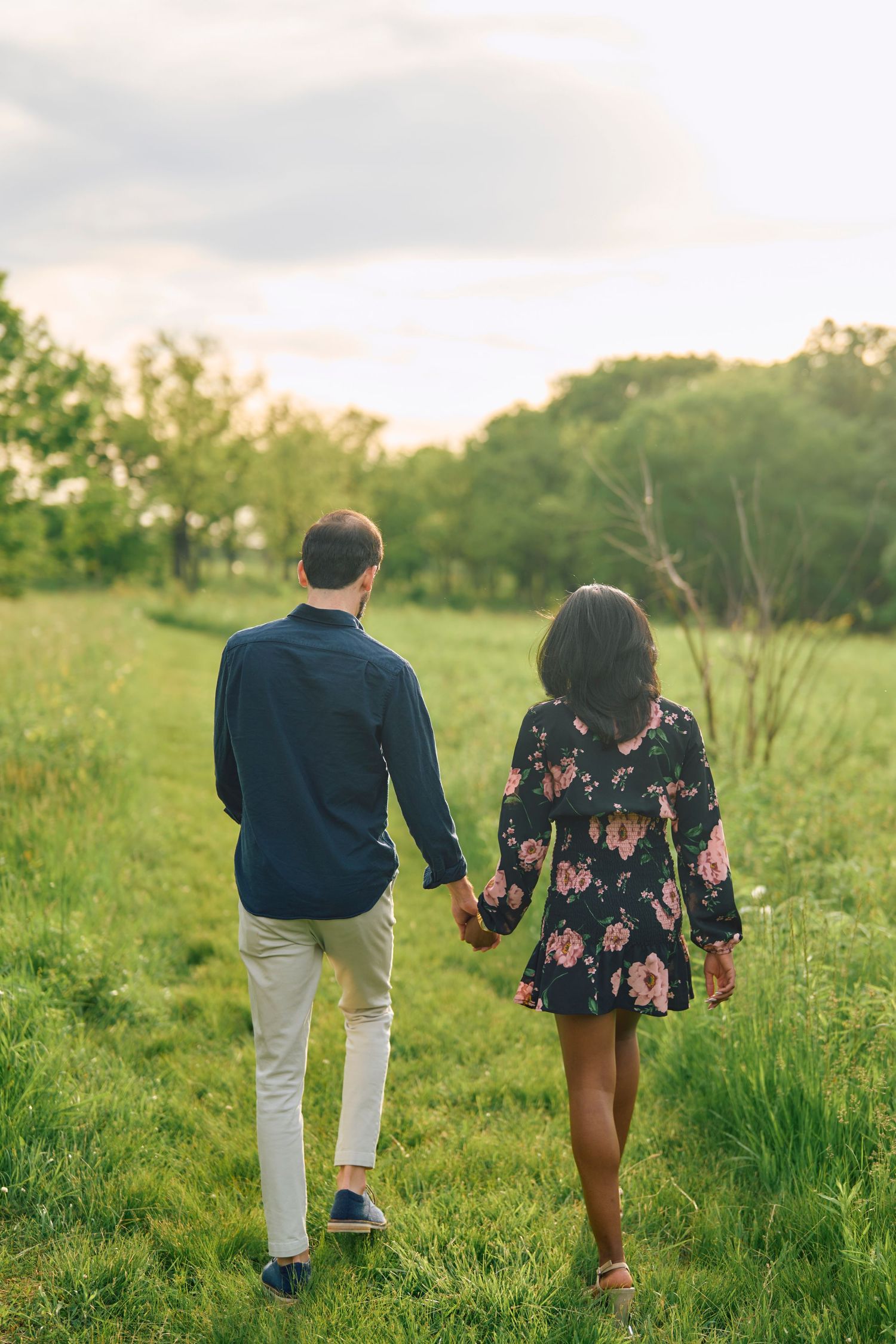 A couple walks hand in hand through a lush green meadow on a warm evening surrounded by trees.