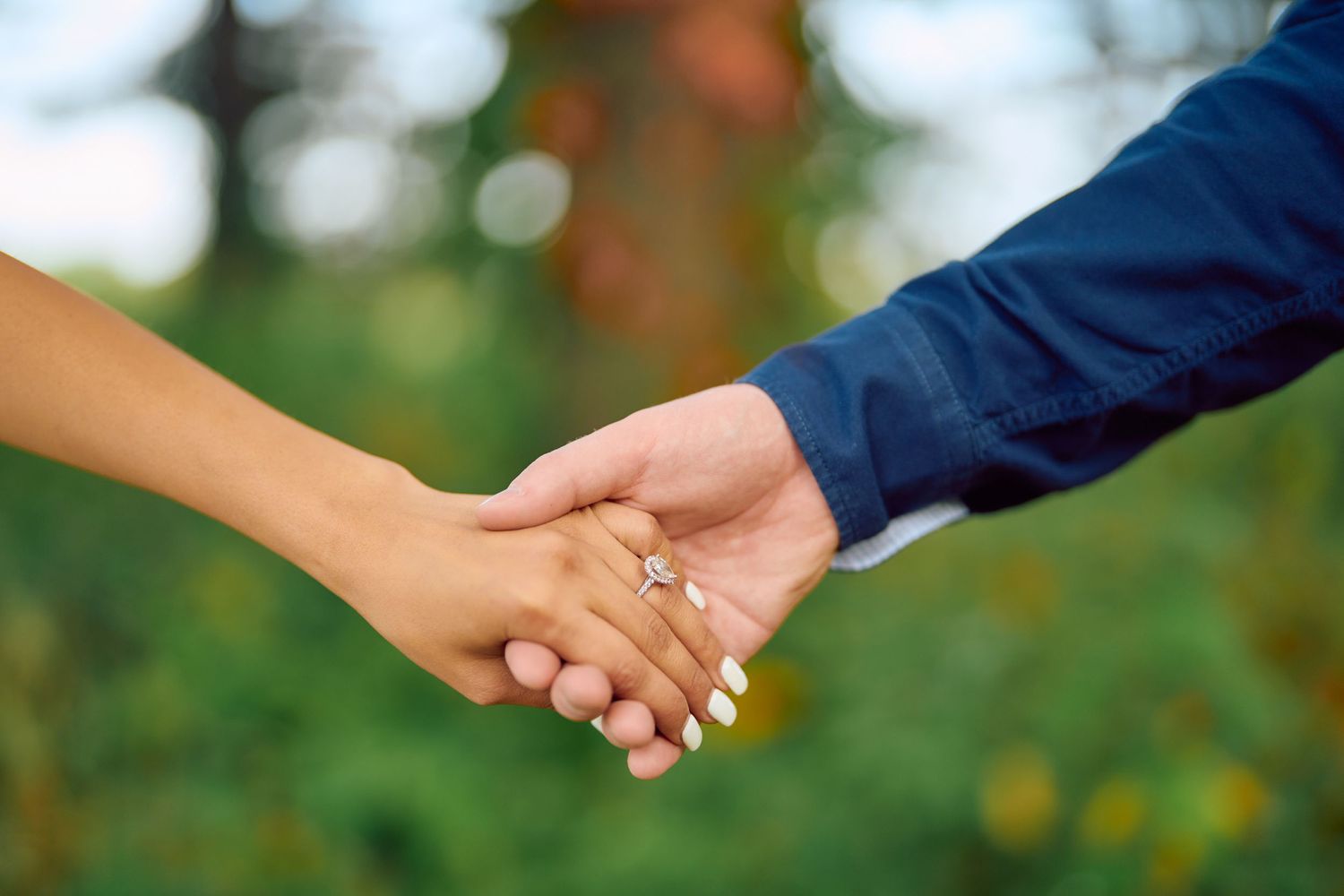 Close-up shot of a couple's hands holding together with an engagement ring, against a blurred green outdoor background.