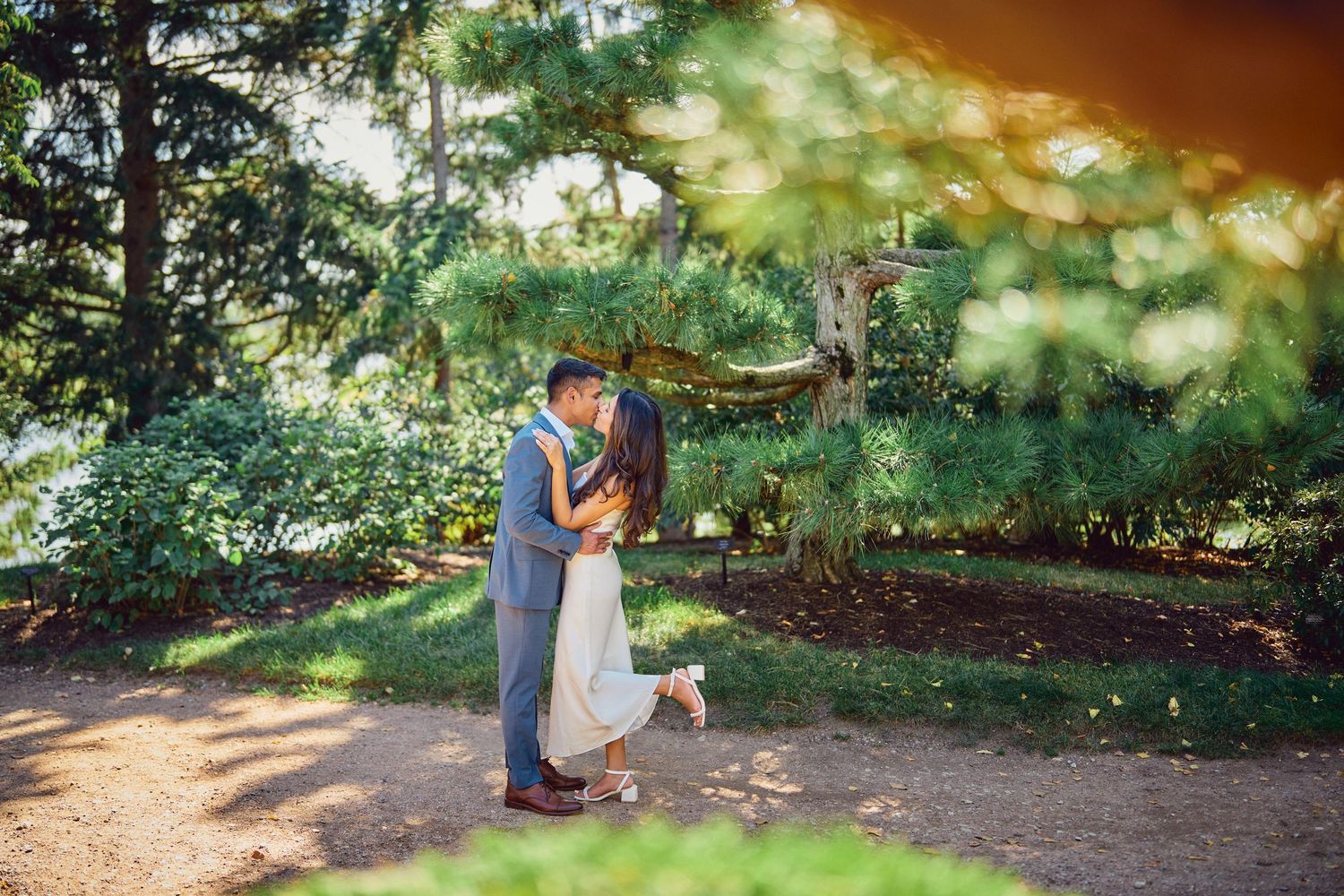 Two people share an intimate moment in a sunlit garden path surrounded by lush greenery.