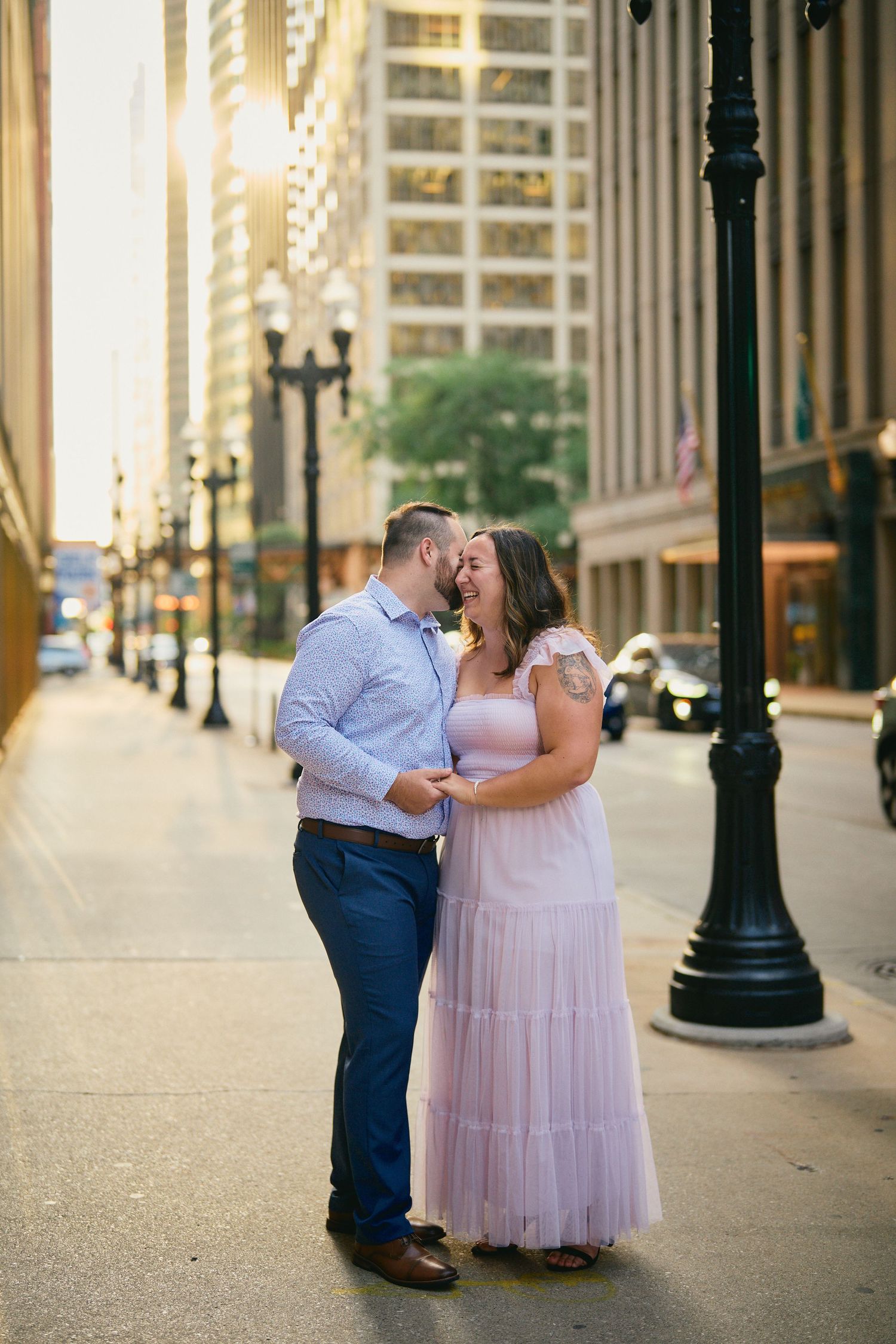A couple shares an intimate moment on a city street with lamp posts and tall buildings in golden sunlight.