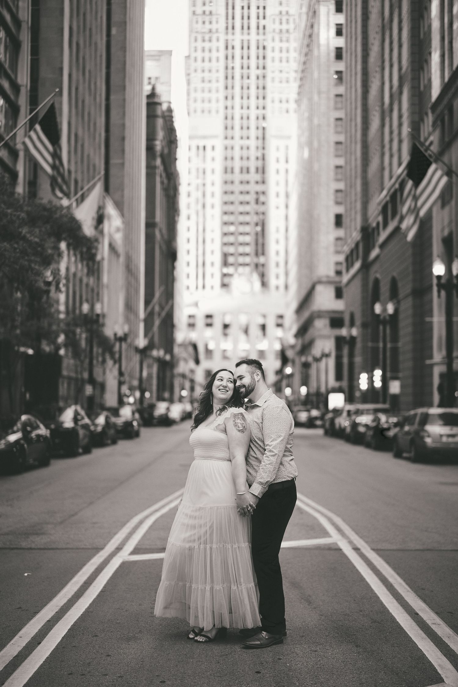 A romantic black and white urban photo featuring a couple standing in the middle of a Chicago street with tall buildings.