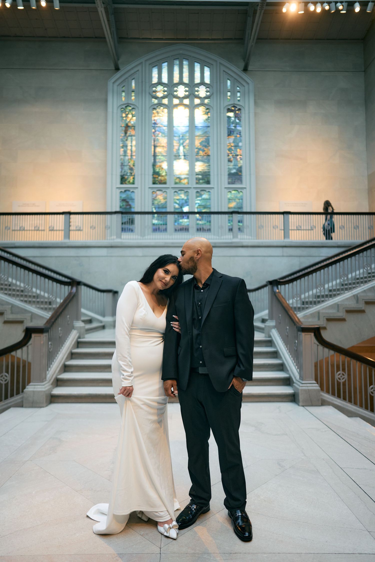 Elegant couple poses on grand staircase with ornate stained glass window in background at formal venue.