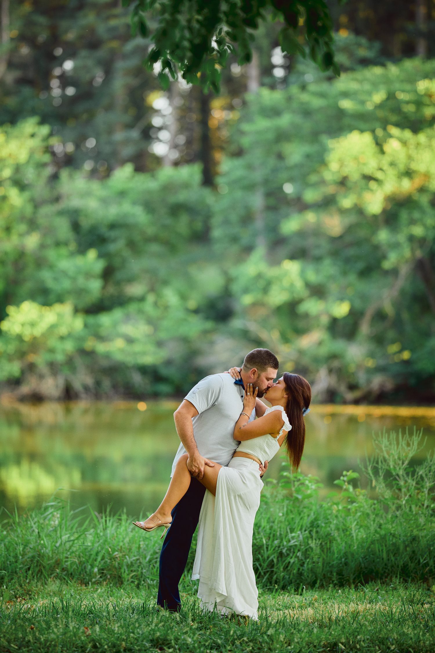 Romantic couple embraces in an intimate pose on grassy lawn beside a tranquil pond surrounded by lush summer foliage.