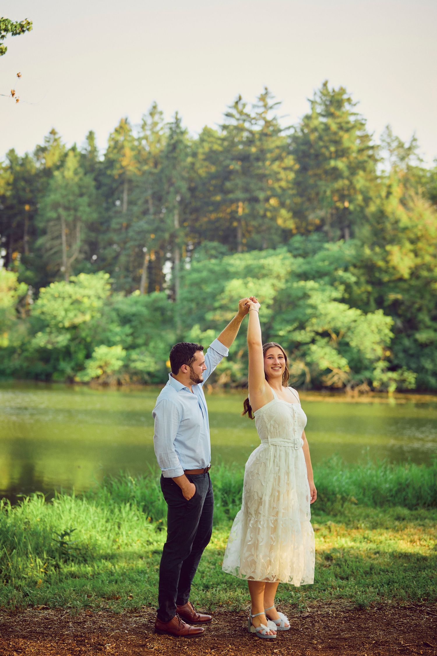 A couple holds hands and dances by a serene lake with evergreen trees in the background during golden hour.