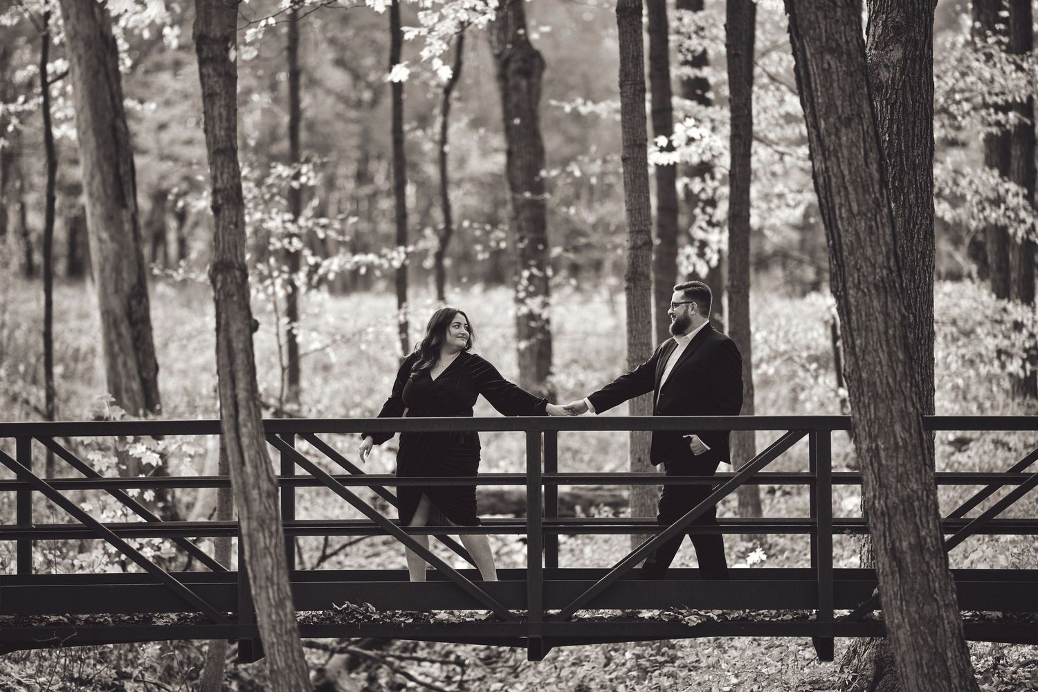 A romantic black and white photograph of two people sharing a moment on a wooden bridge in a wooded setting.
