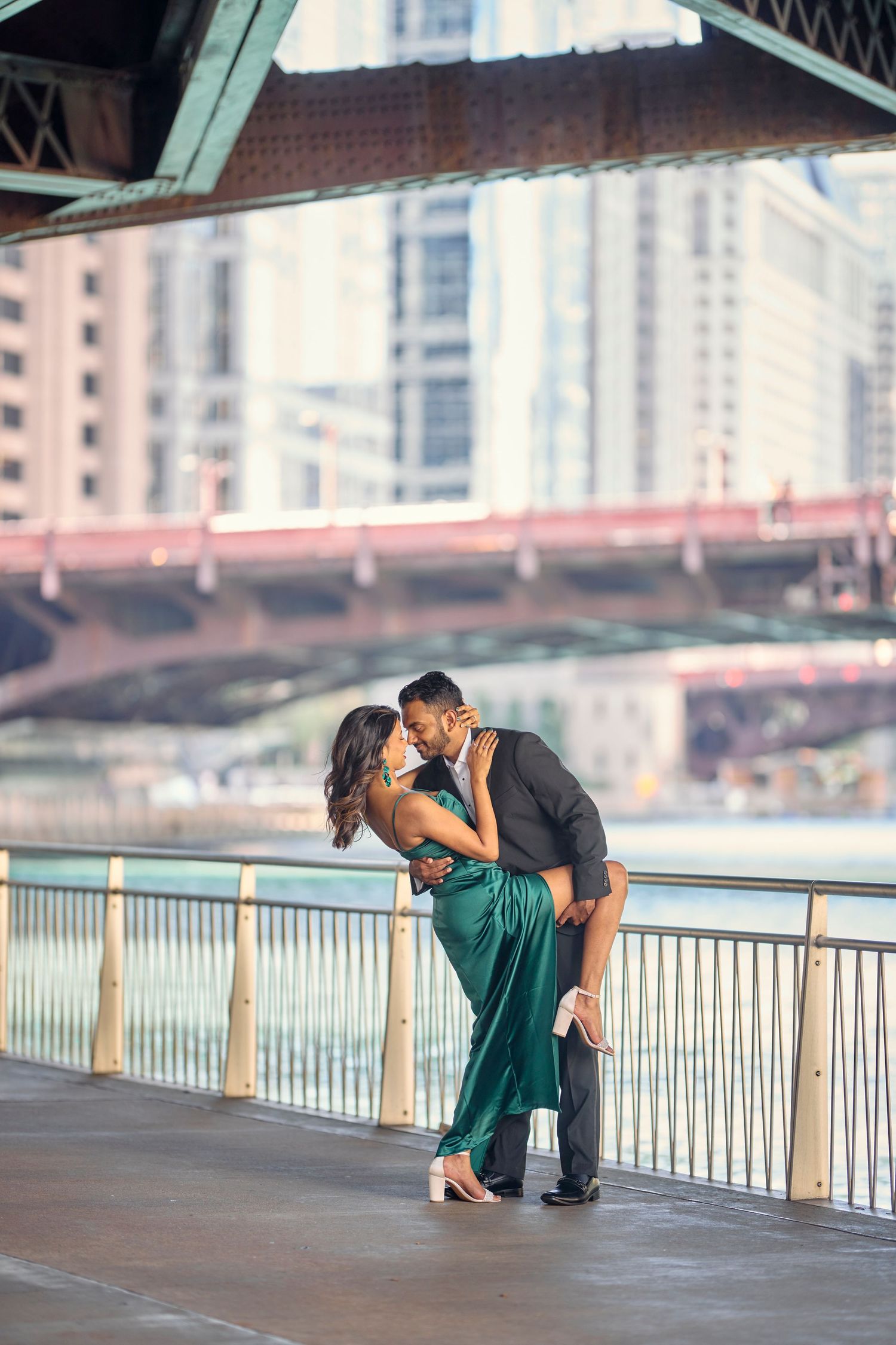 A romantic couple embraces on a bridge in Chicago with the city skyline in the background and greenposts along railing.